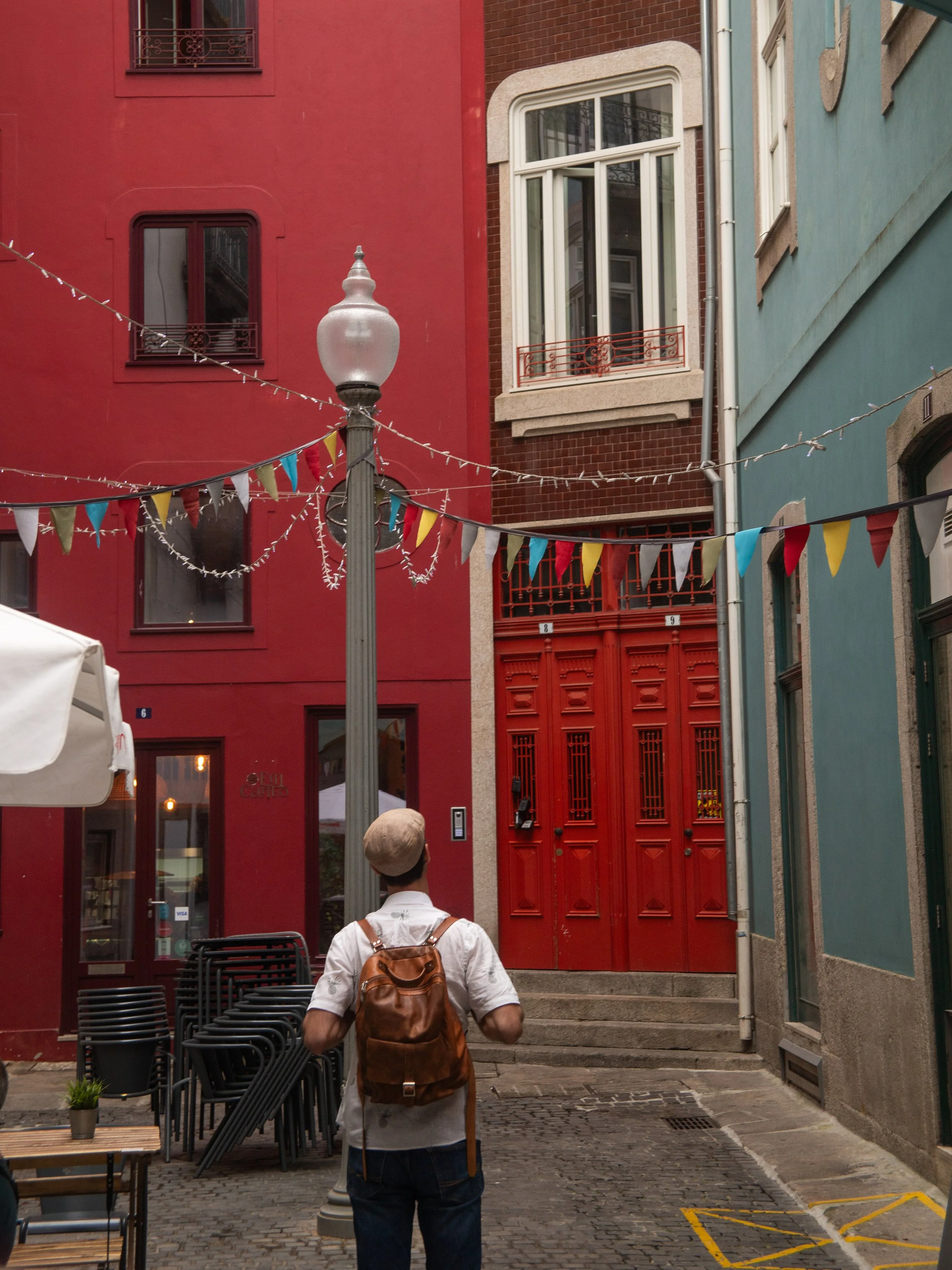 A person with a backpack and hat standing on a cobblestone street, looking toward a red door in between colorful buildings, with decorative string lights and bunting overhead.