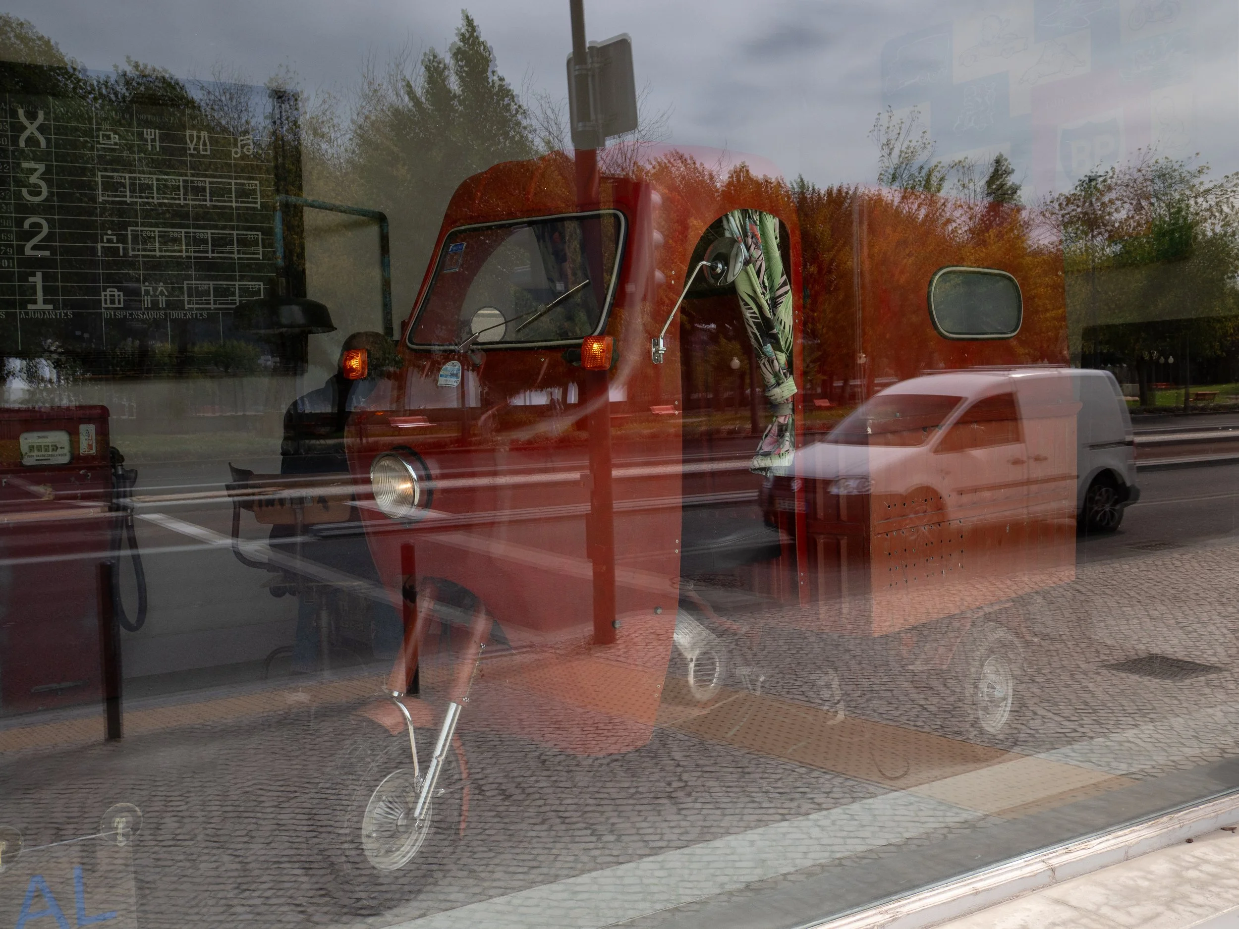 Reflection of a red three-wheeled utility vehicle and a white van in a glass window, with trees and a street visible in the background.