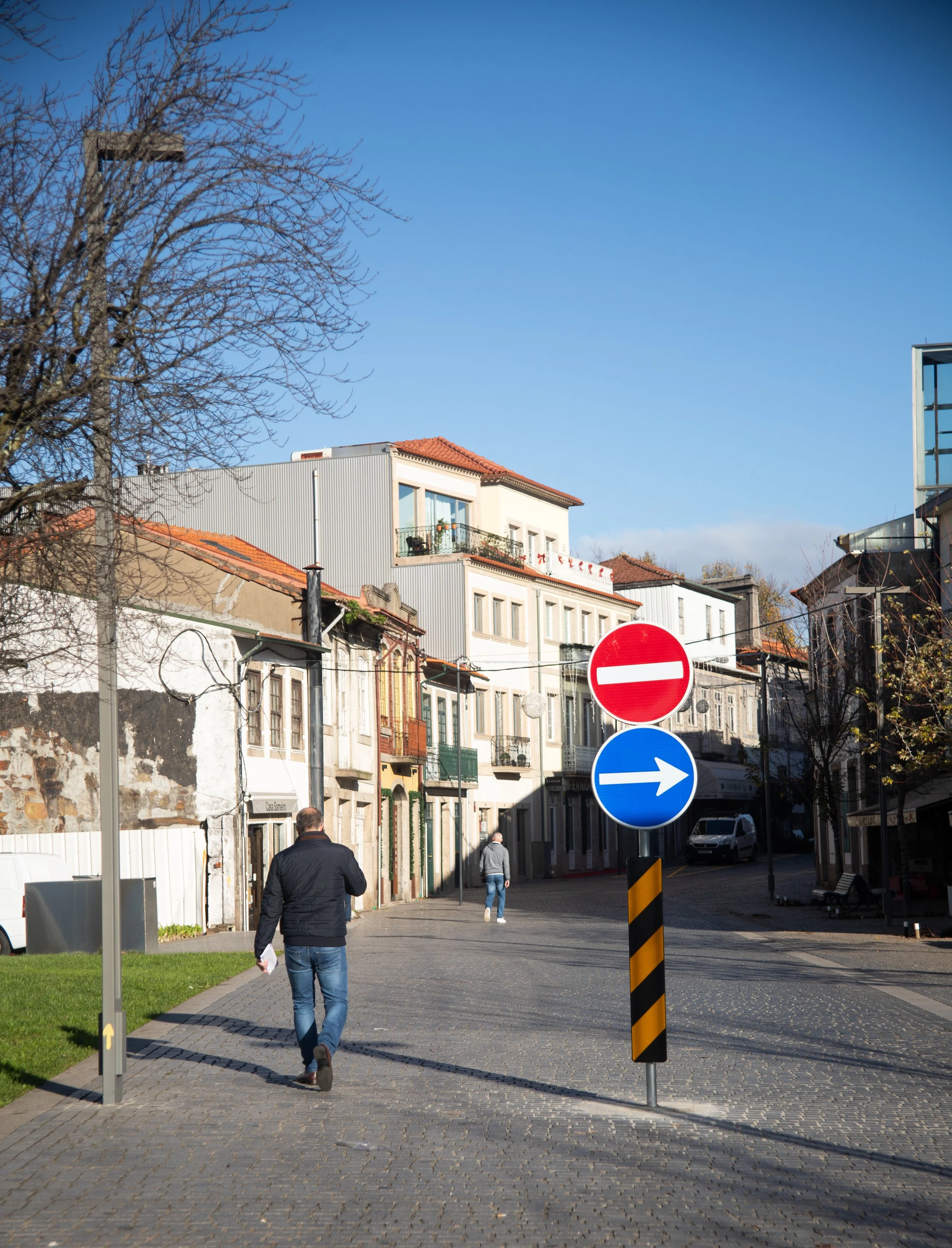 Street scene with two pedestrians walking, street signs indicating no entry for vehicles from the left and a direction to the right, buildings with mixed architecture, a leafless tree, and a clear blue sky.