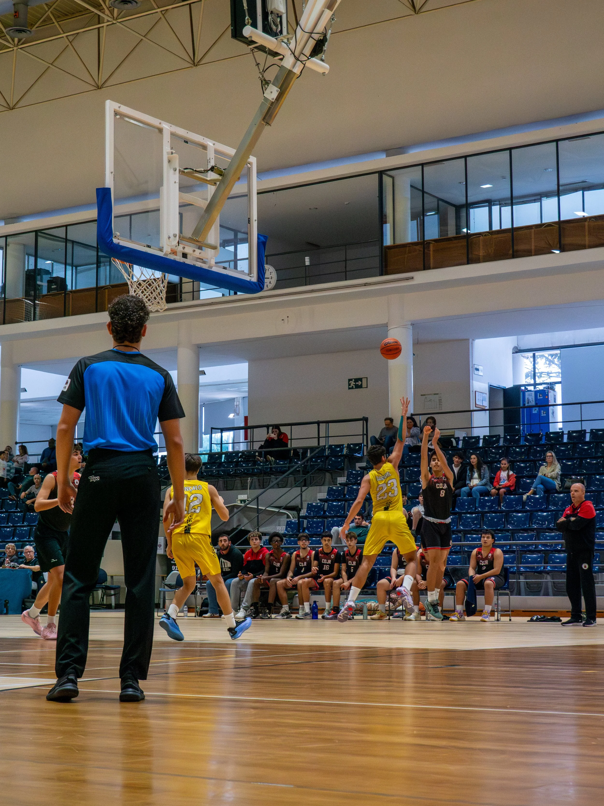 A basketball game in an indoor gym, with players in yellow and black uniforms. One player in black is jumping to shoot or block, while a player in yellow is attempting to block. Spectators are seated in the background.