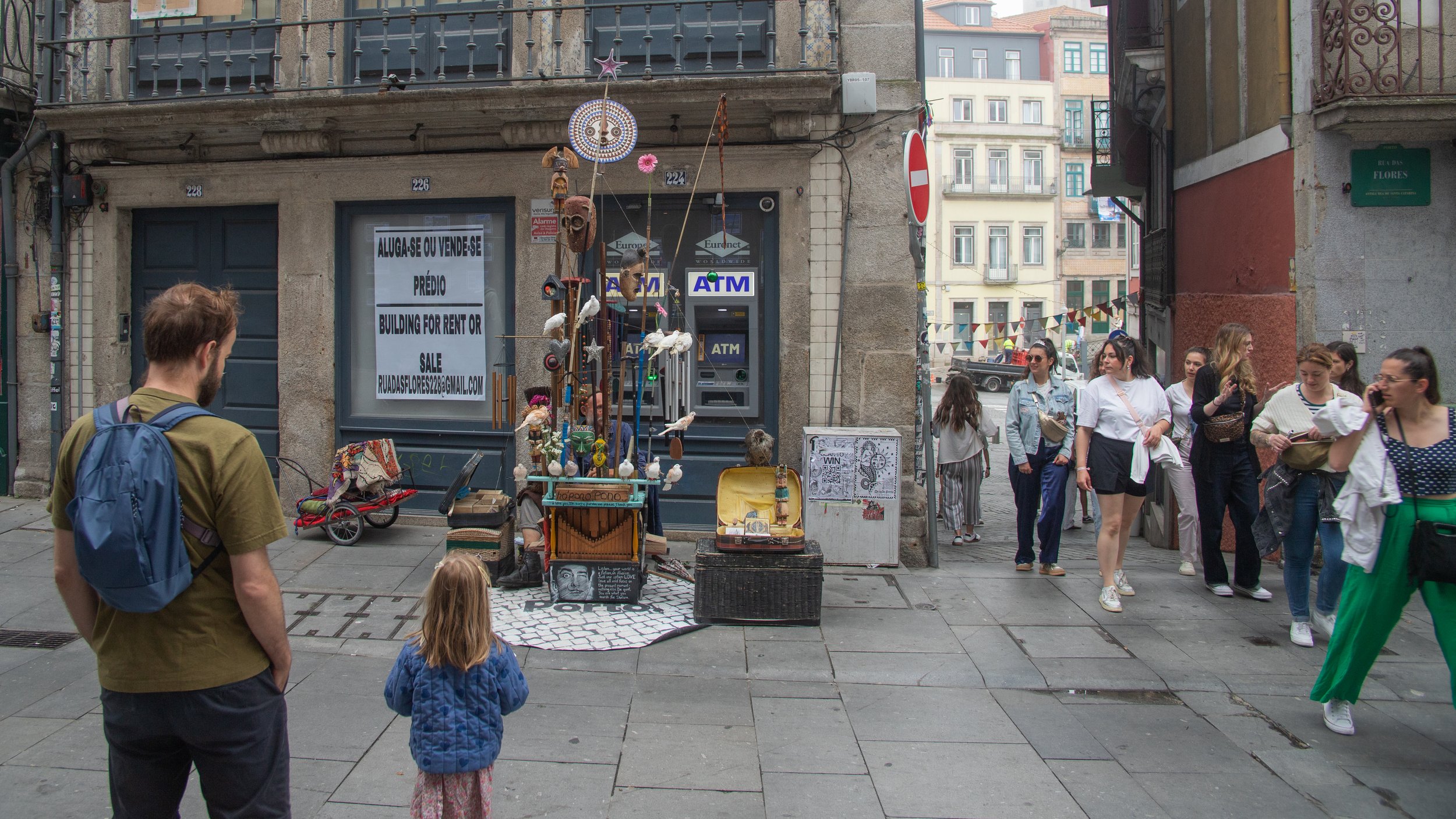 A street scene featuring a group of pedestrians walking along a sidewalk in an urban area. There is a street vendor or street performer with a display of handcrafted items and decor in front of a building. People are casually dressed, some are on the