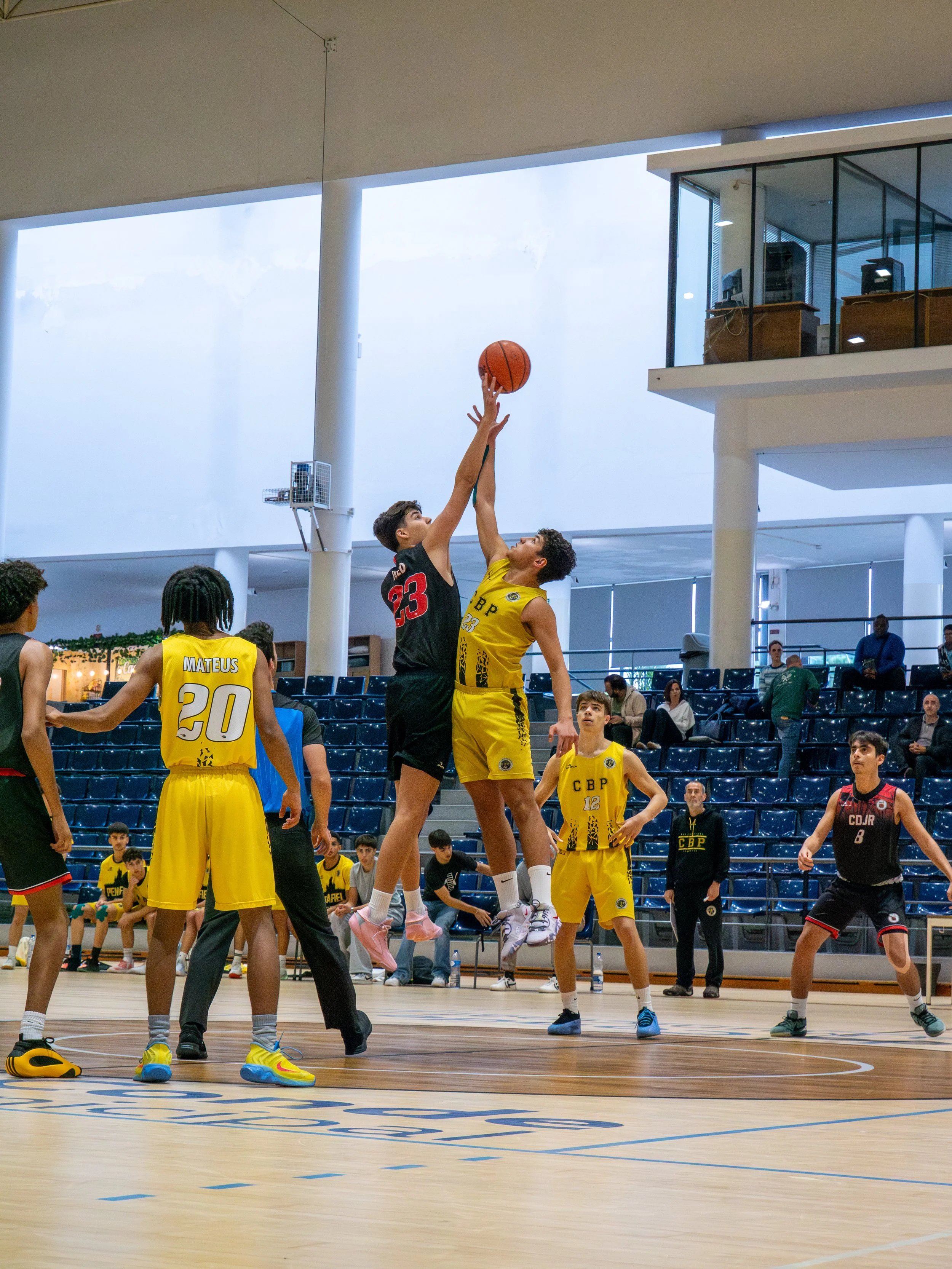Two young male basketball players jump to reach the ball at the tip-off, one in a black jersey and the other in a yellow jersey, during a game in an indoor basketball court. Several other players and spectators are visible in the background.