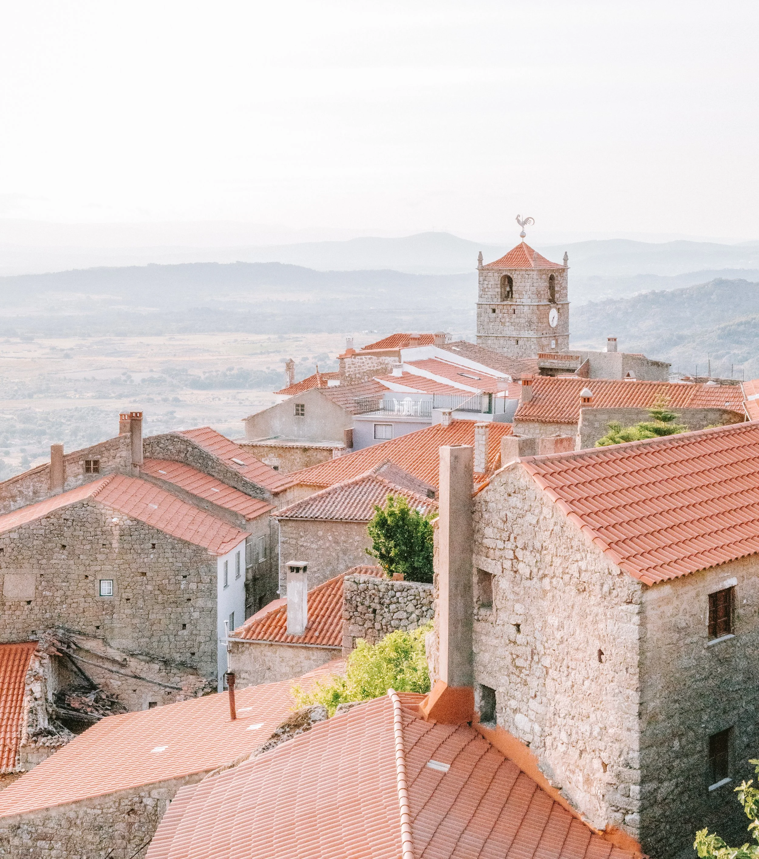 Scenic view of a small European village with stone buildings and red-tiled roofs, a clock tower with a weather vane, and distant rolling hills.