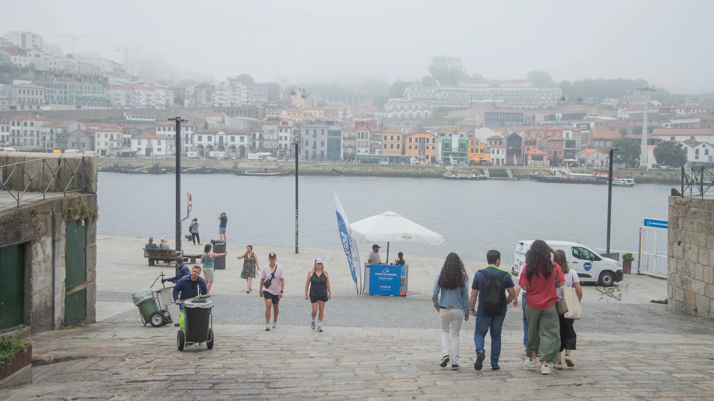 View of a riverfront with people walking down stone steps, some sitting on benches, and colorful buildings on the hillside across the water, with fog partially obscuring the background.