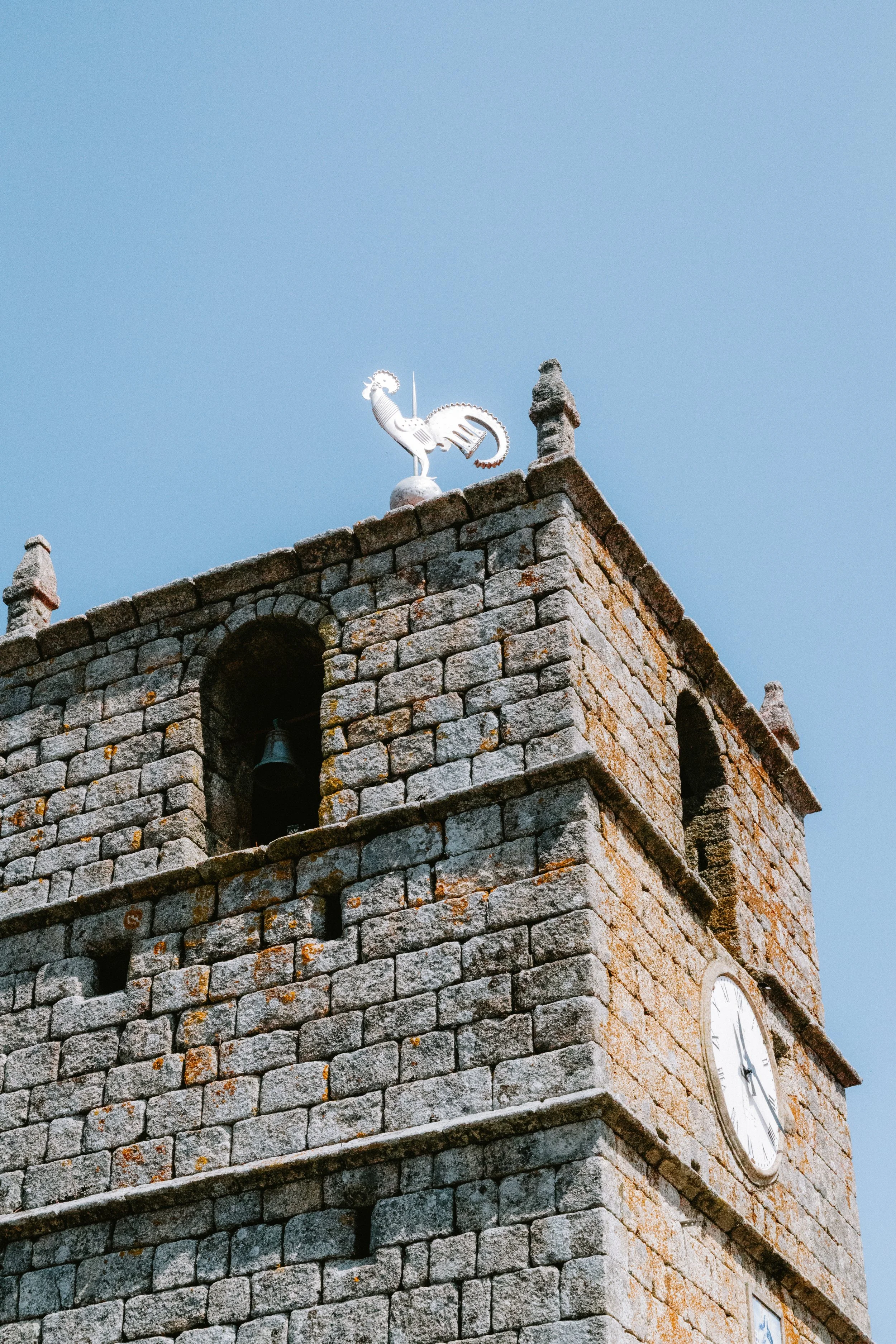 Close-up of a stone clock tower topped with a metallic weather vane shaped like a rooster, against a clear blue sky.