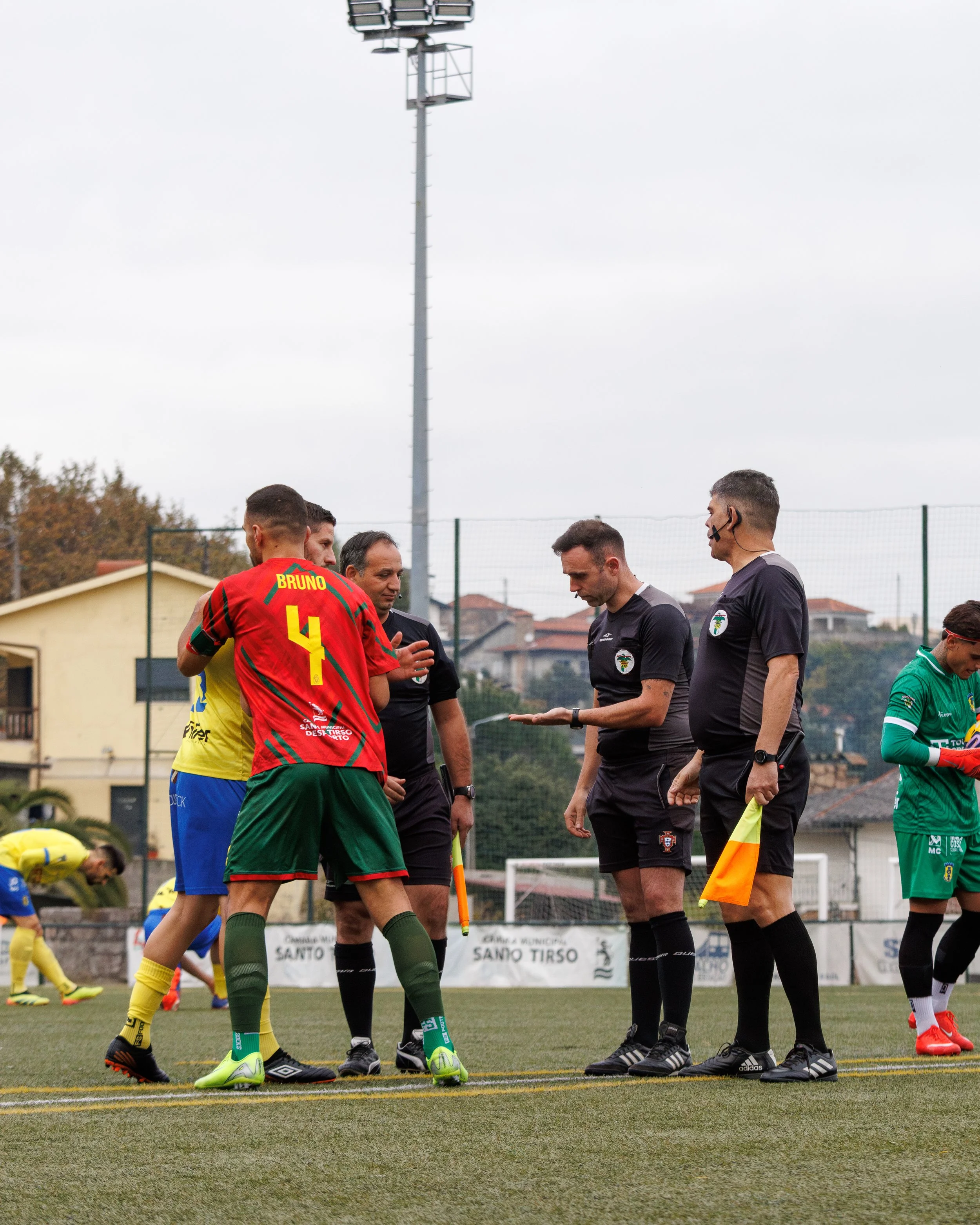 Soccer referees and players preparing for a match on a soccer field, with a captain in a red and green uniform and a coach in yellow, during daytime with an overcast sky.