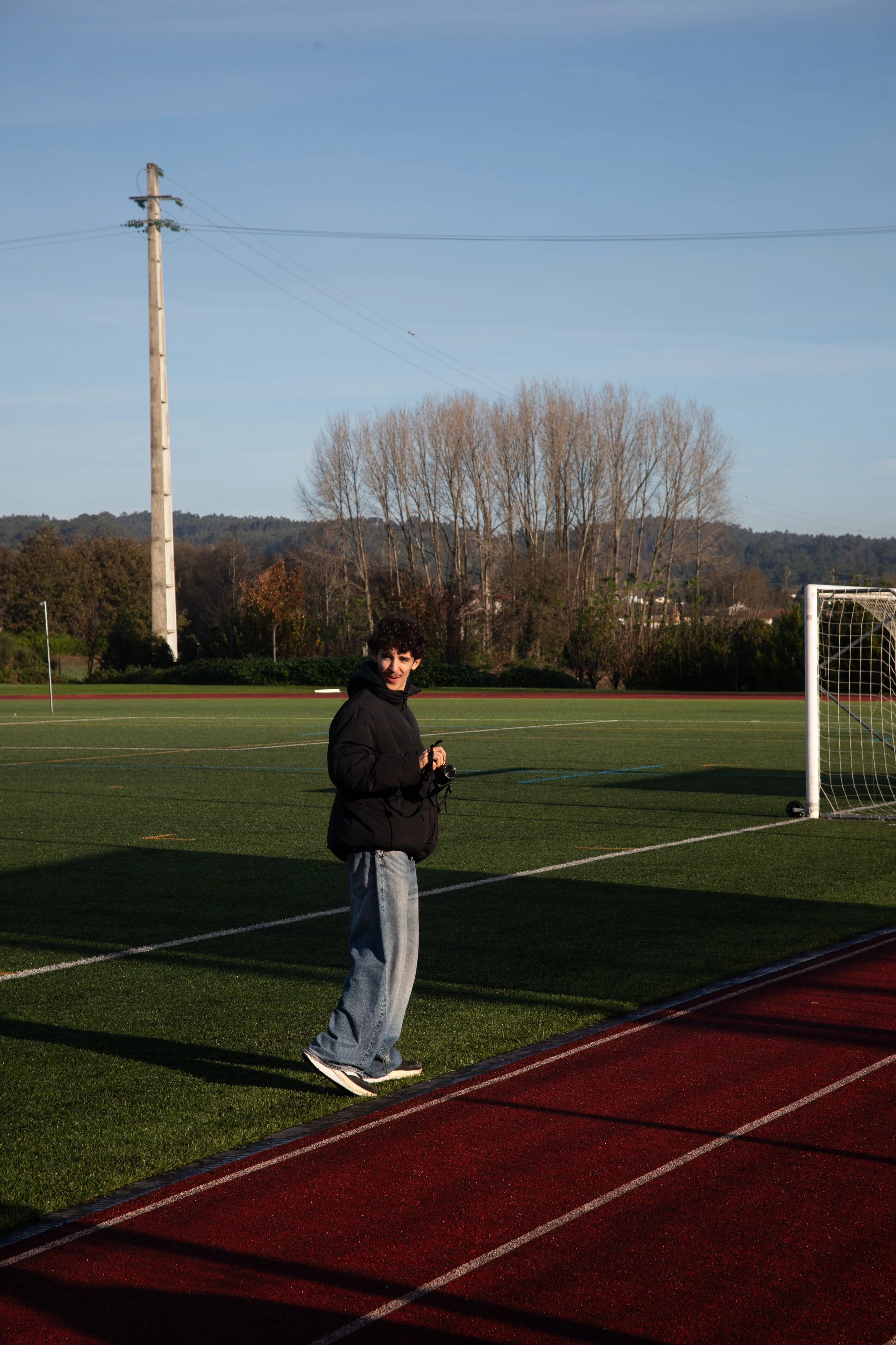 A young man in a black jacket and jeans standing on a running track next to a grass soccer field, holding a camera. The scene is outdoors with clear blue skies, leafless trees, and a power pole in the background.