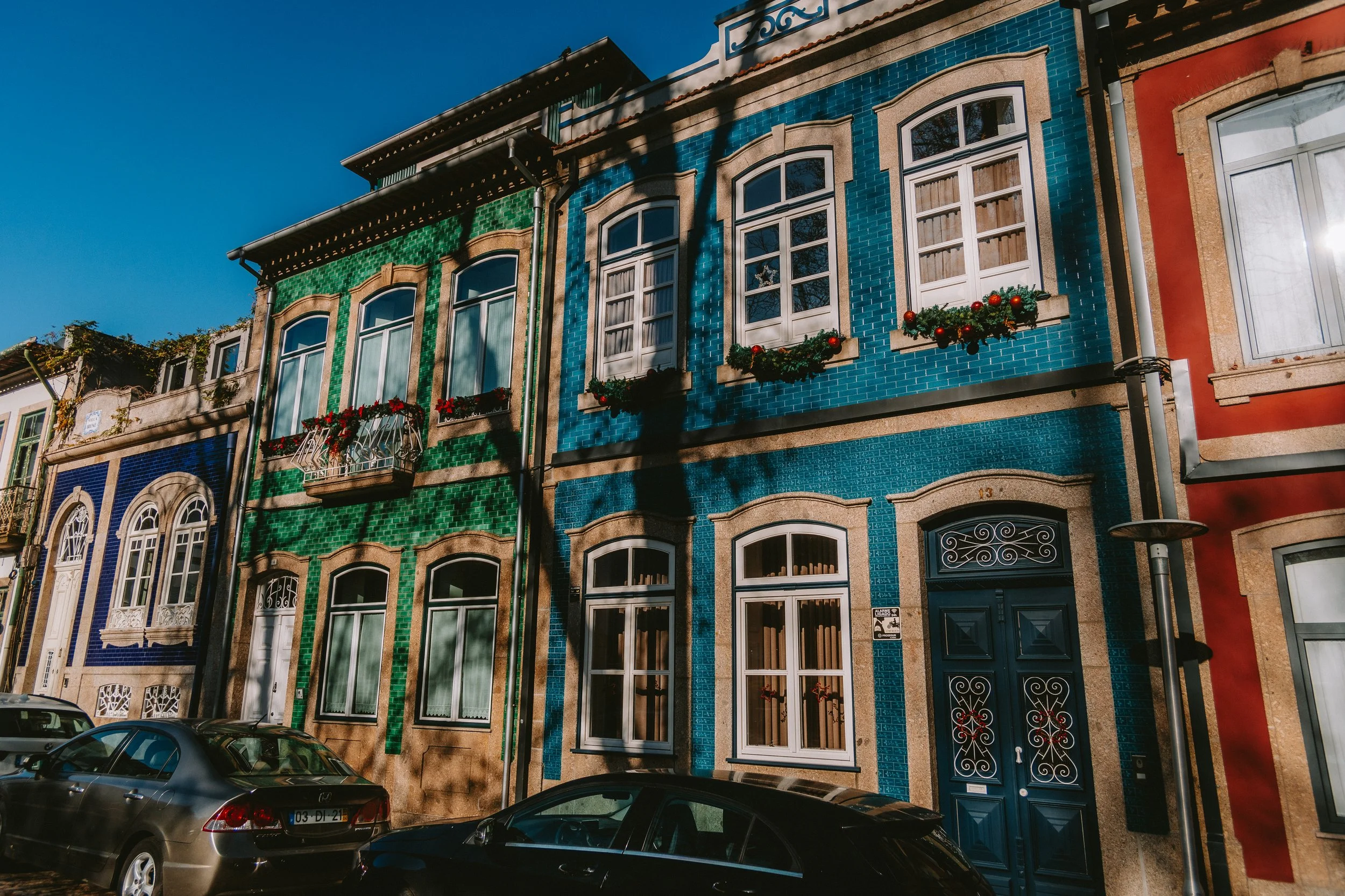 Colorful old two-story building with blue and green tile facades, decorated with Christmas ornaments and flowers in window boxes, parked cars in front, under a clear blue sky.