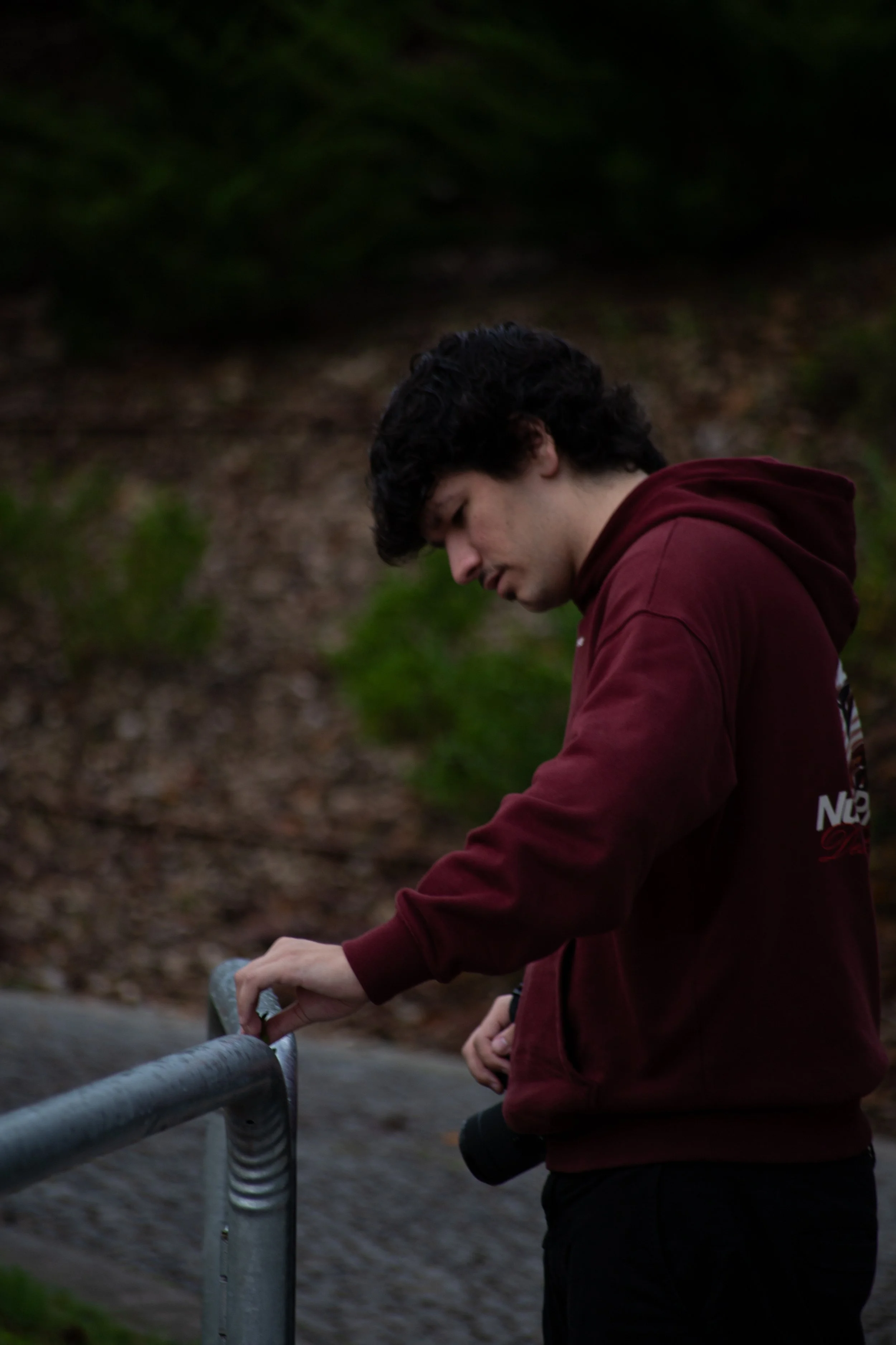 Young man with curly dark hair, wearing a maroon hoodie, standing outdoors with a camera hanging from his belt, looking down and touching a metal railing.