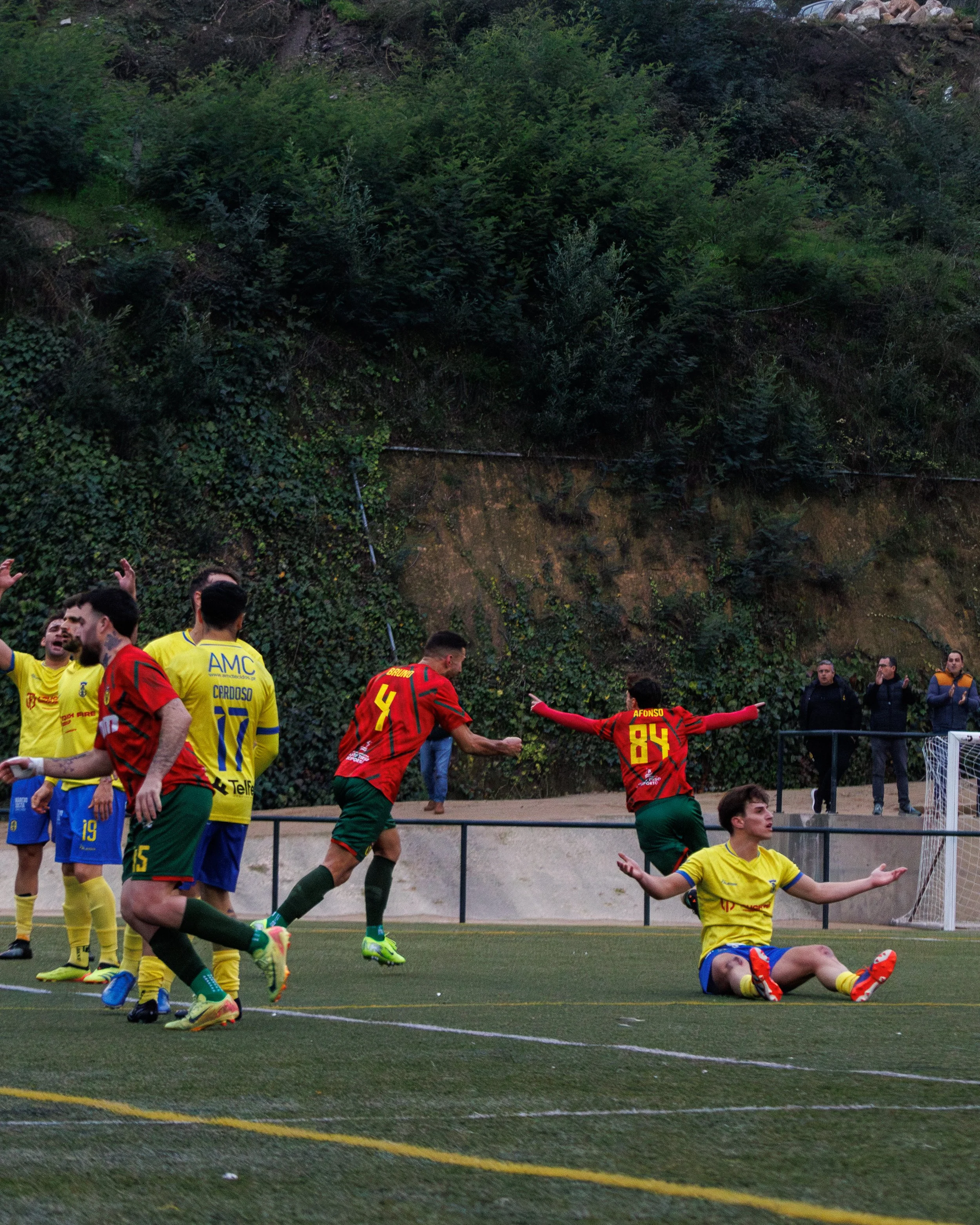 Soccer match with players celebrating and one player sitting on the ground, in front of a green hillside.