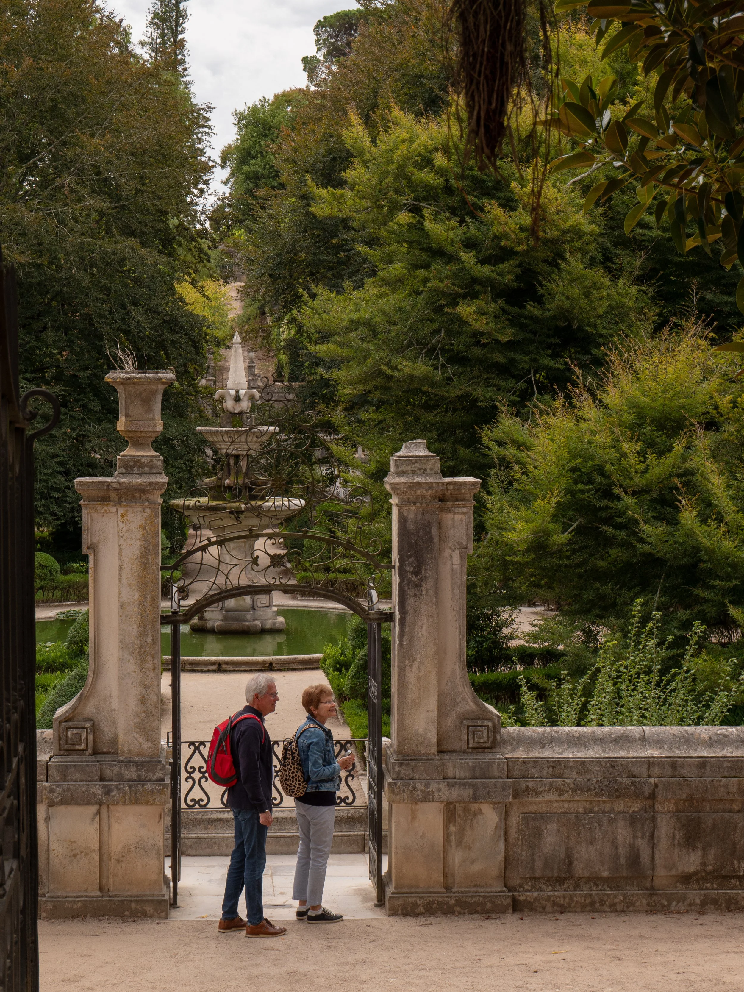 Older man and woman with backpacks standing at an ornate iron gate in a lush garden, engaging in conversation. The background features a stone statue in a pond and a sloping path lined with dense greenery.