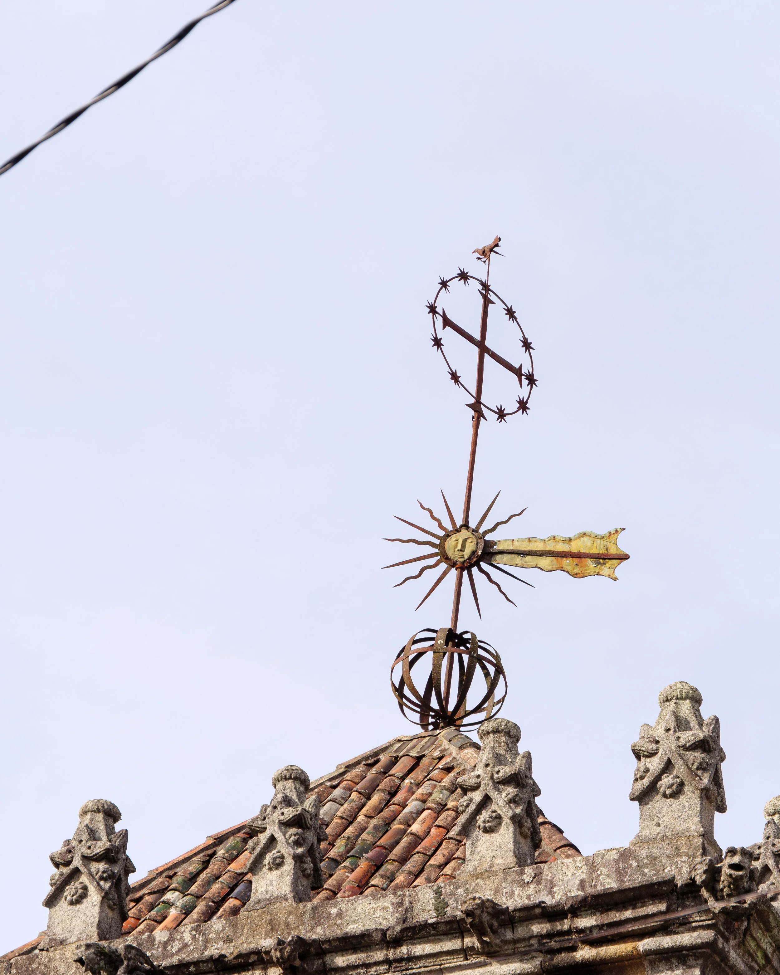 An ornate iron cross with a sun design and star-shaped circle, mounted on a tiled roof with decorative stone carvings, against a pale blue sky.