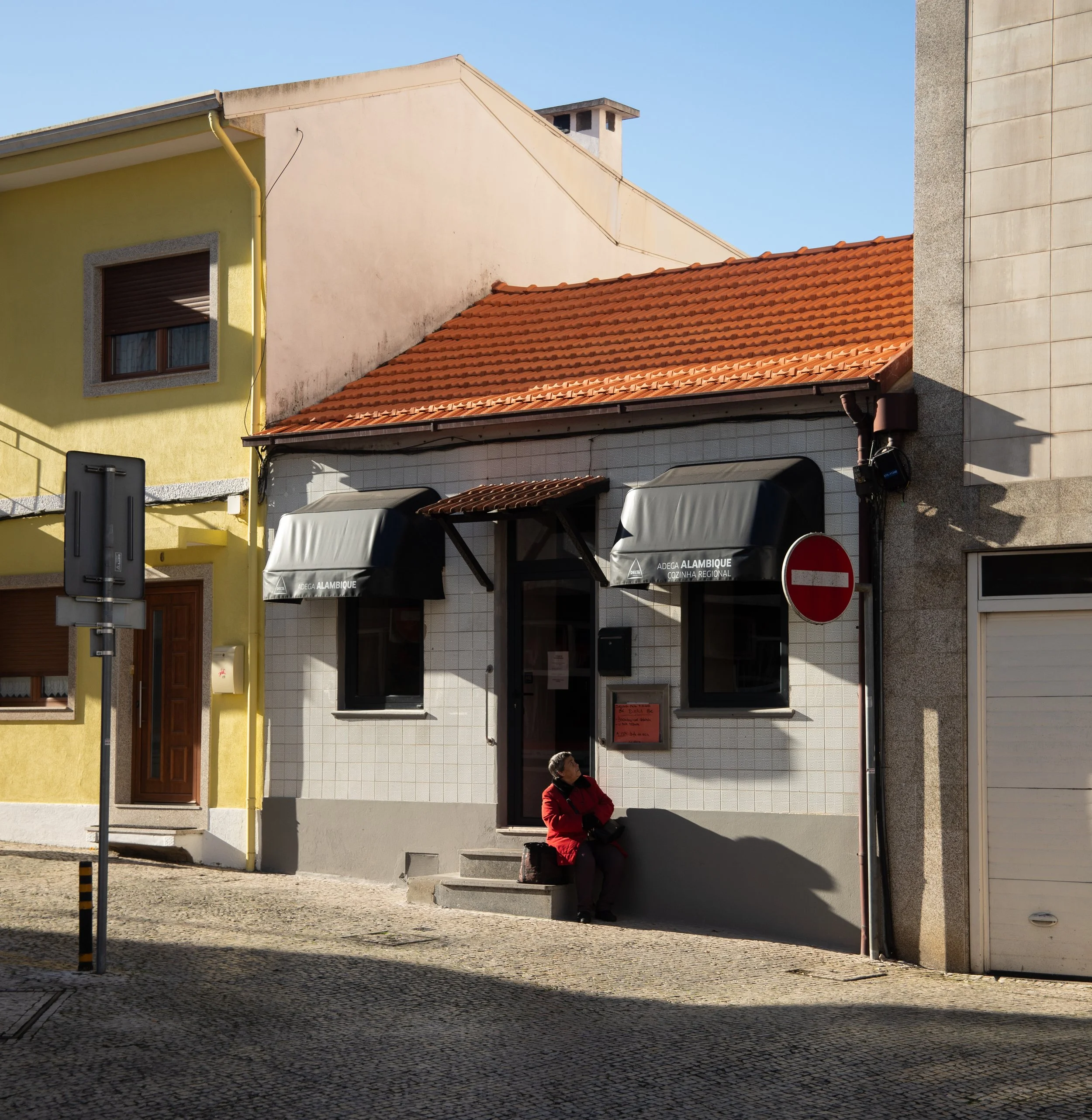 A woman in a red jacket sitting on steps outside a small restaurant with black awnings, on a cobblestone street in a European town.