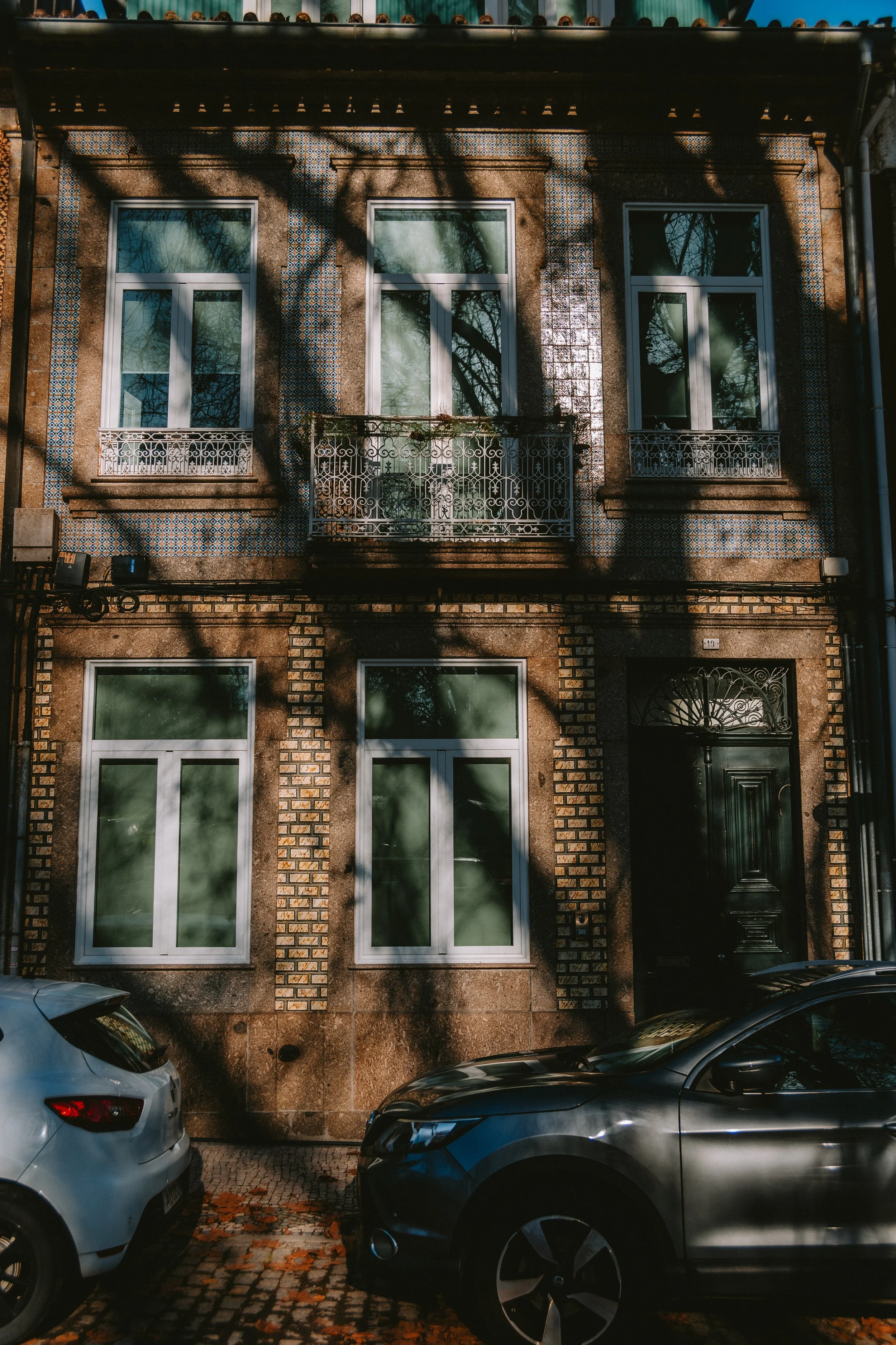 Three-story brick residential building with four windows, two on each floor, and a decorative black front door. Shadows from tree branches are cast on the facade. Two cars are parked in front, one white and one dark gray.