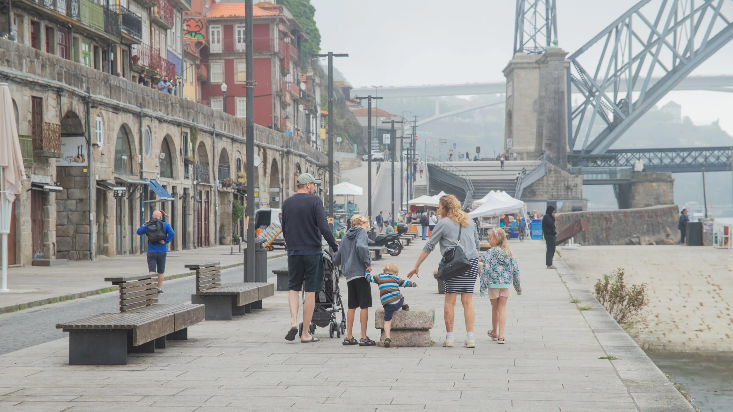 A group of people, including two children and two adults, standing and interacting on a waterfront promenade with benches and lampposts, with colorful buildings, outdoor cafes, and a bridge in the background.