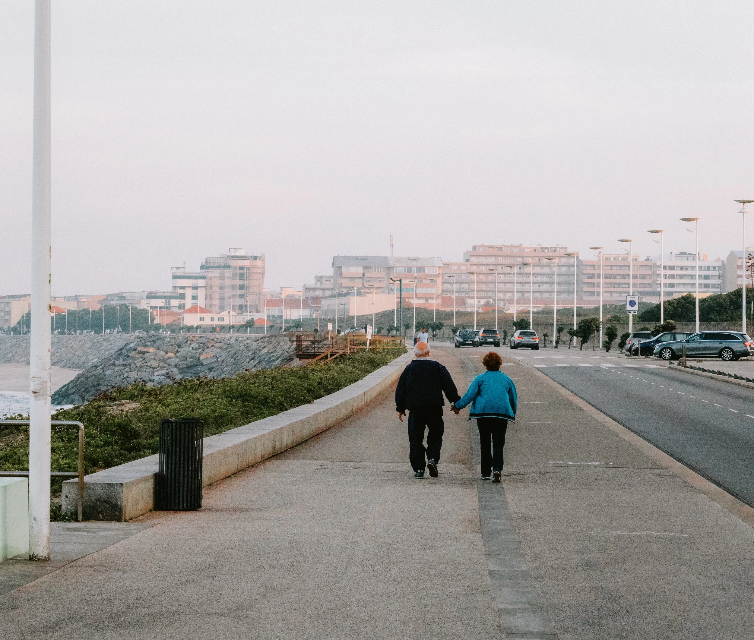 An elderly couple walking hand in hand along a paved walkway near the coast, with modern buildings and street lamps in the background during early evening or dawn.