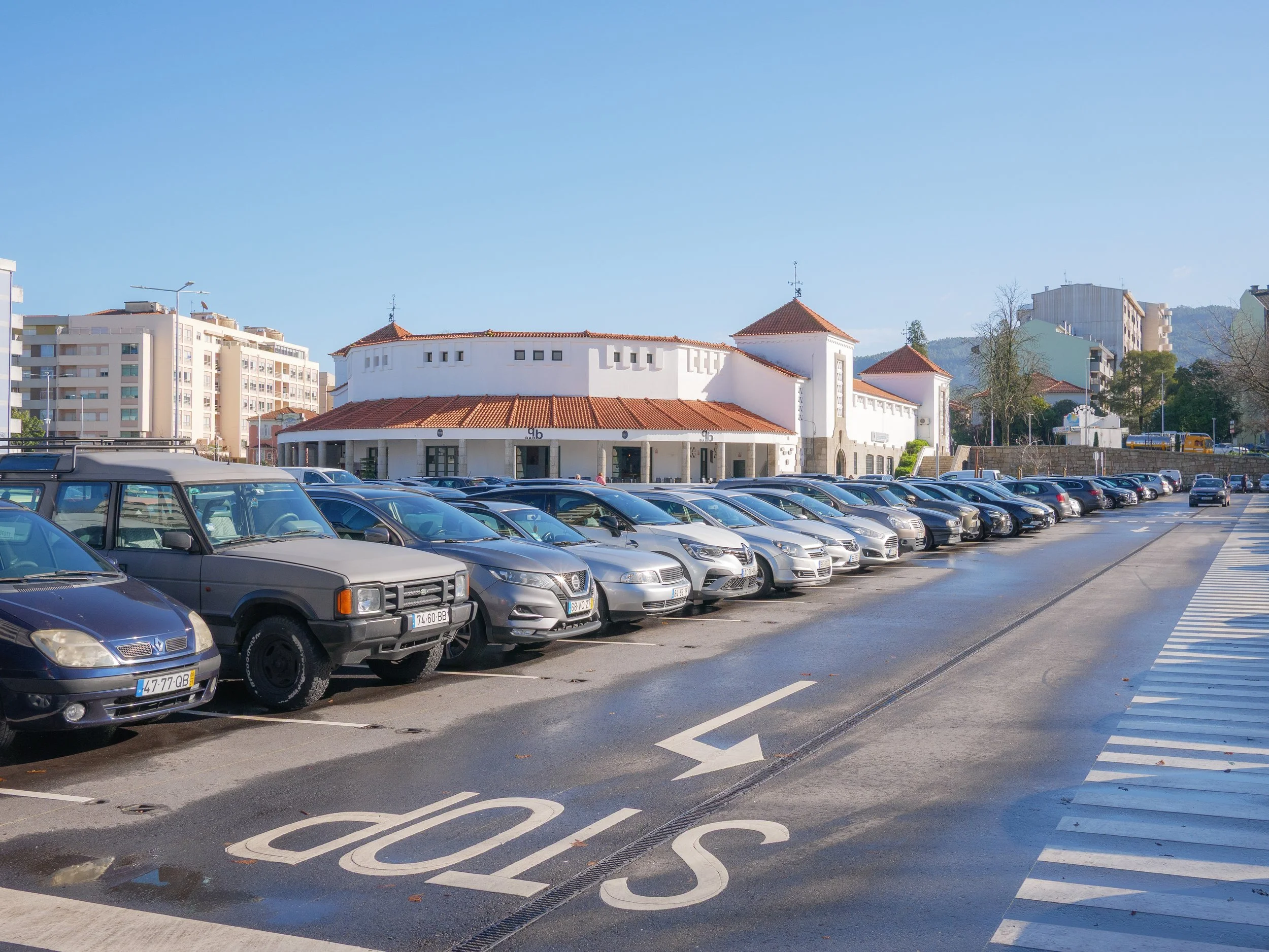 A parking lot with multiple cars parked and a white building with a red-tiled roof in the background.