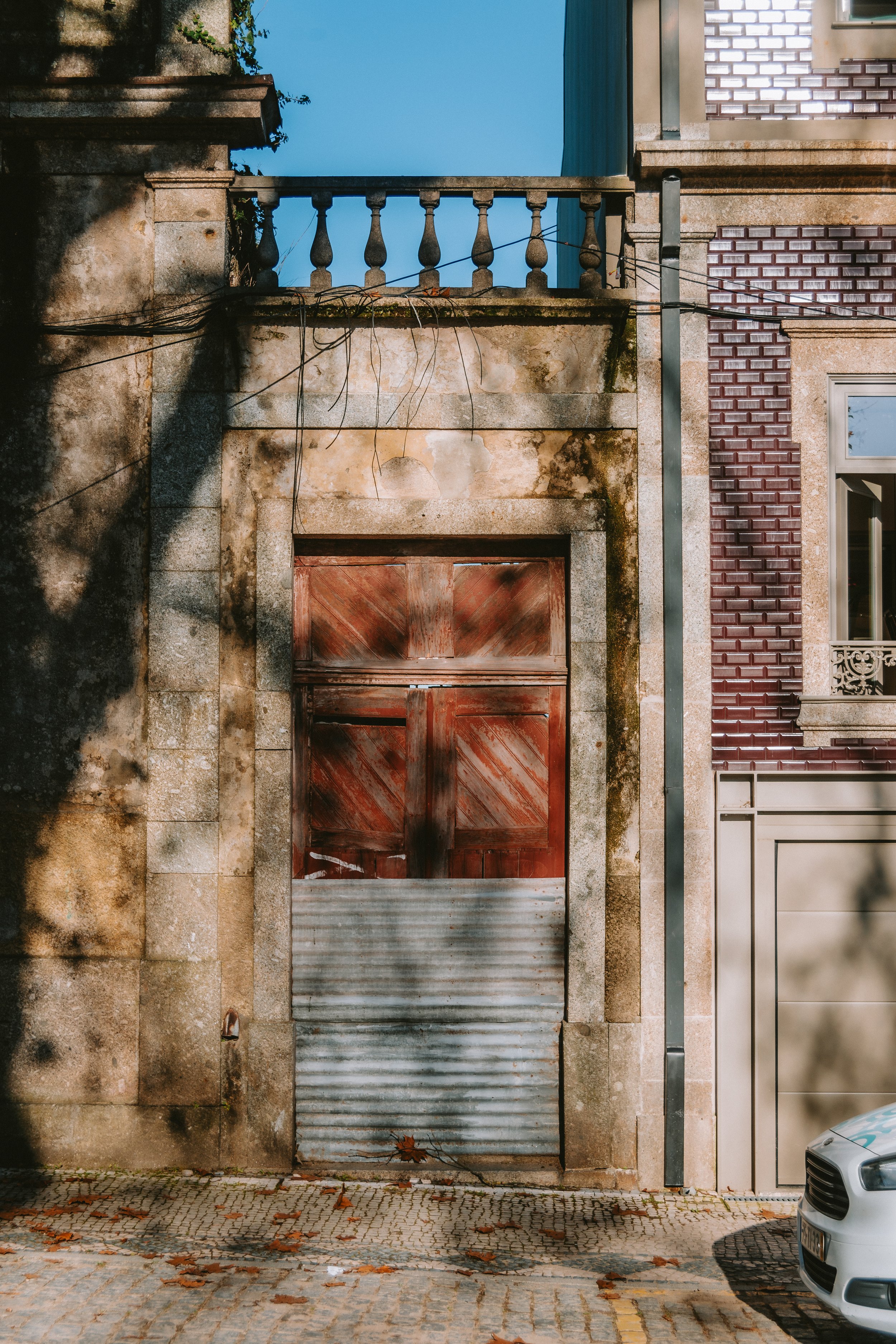 Old wooden garage door with a corrugated metal lower panel, set in a weathered stone wall, with a car partially visible on the right and an ornate balcony above.