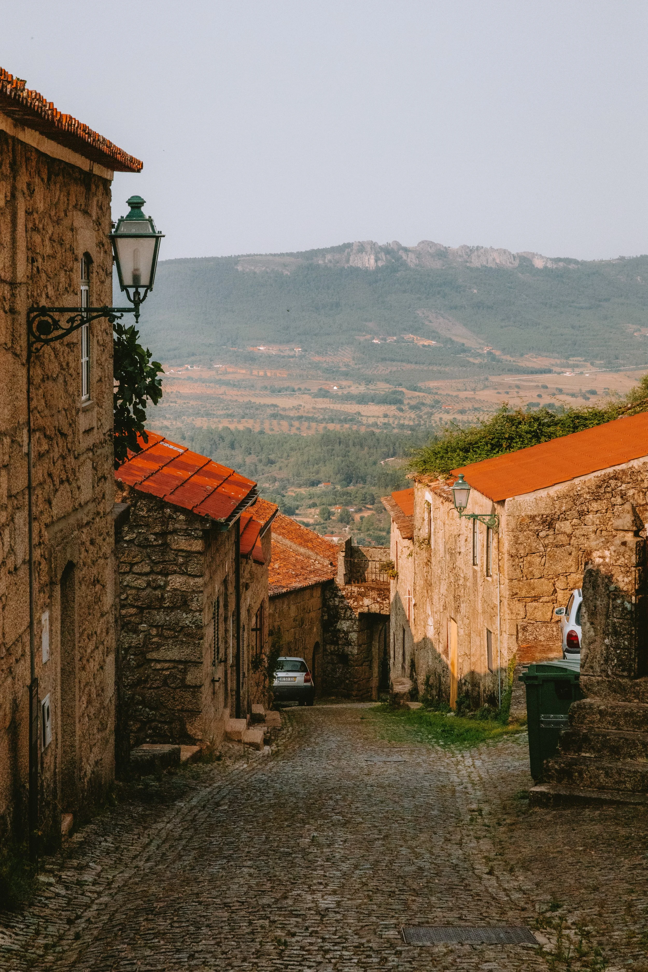 A narrow cobblestone street in a rustic village with stone buildings and terracotta roofs, overlooking a valley with mountains in the background.