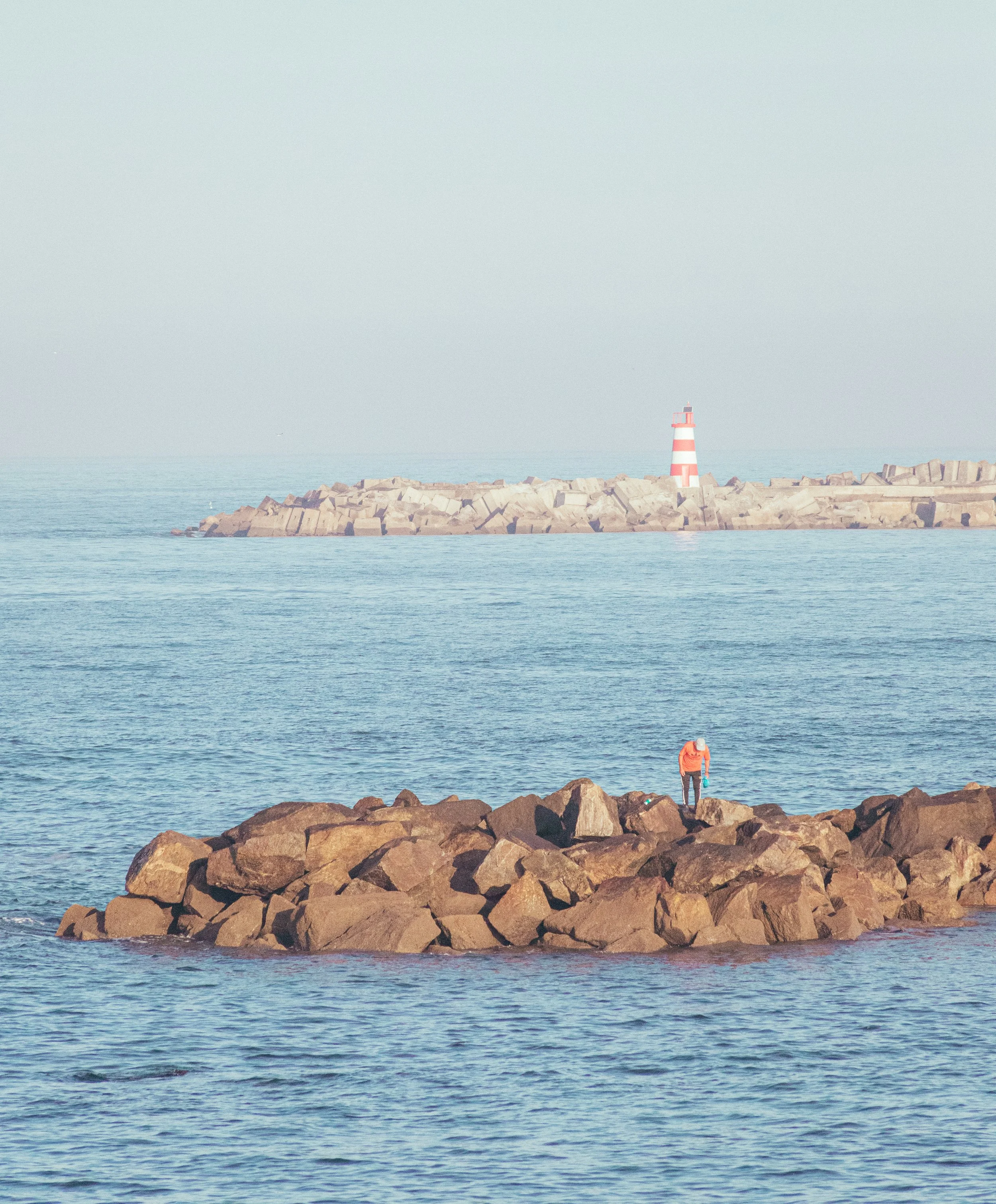 A person standing on a rocky pier by the ocean, with a lighthouse on a breakwater in the background.
