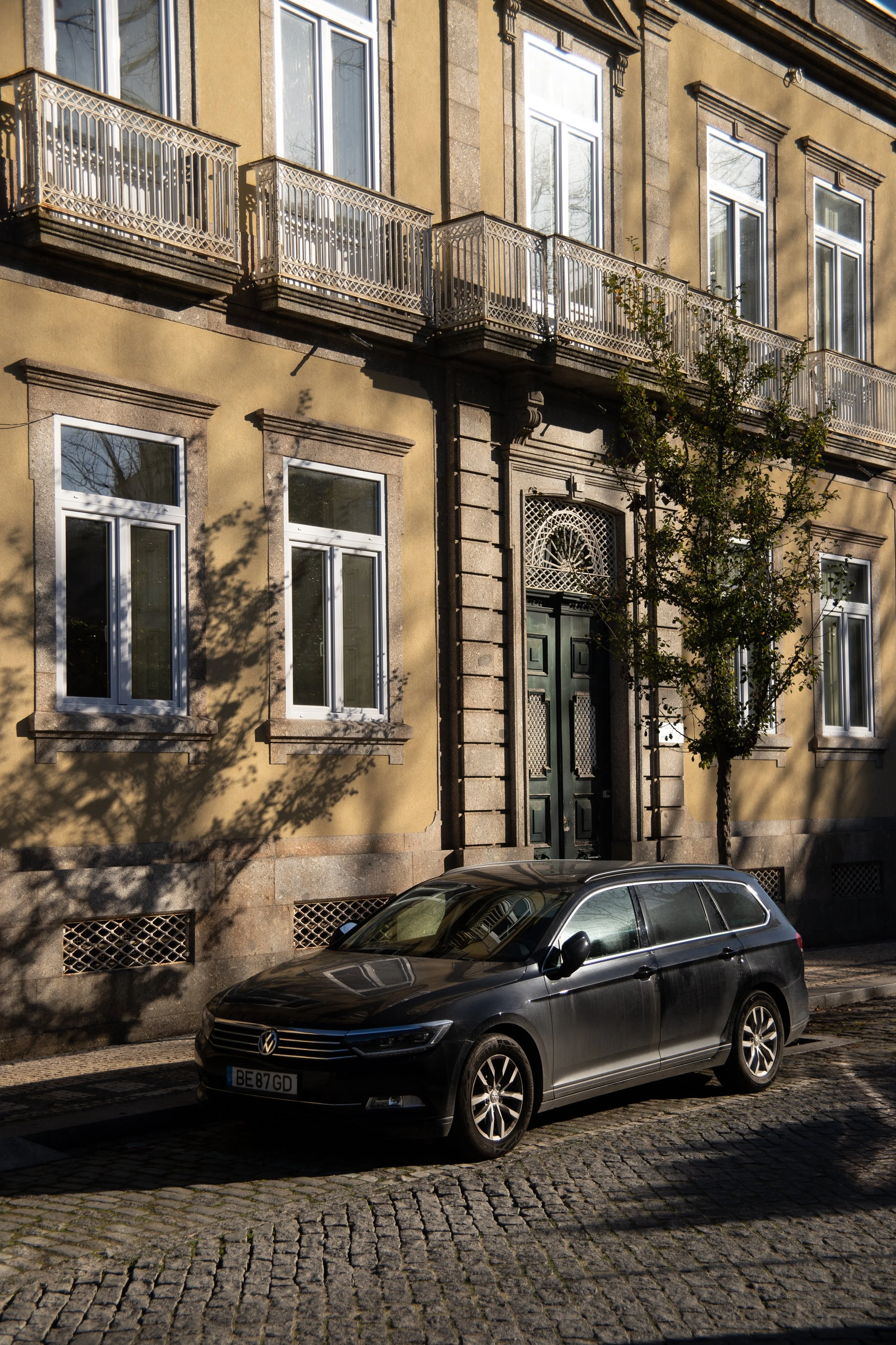 A black Volkswagen car parked on a cobblestone street in front of a beige multi-story building with large windows and ornate entrance door, casting shadows of a nearby tree.