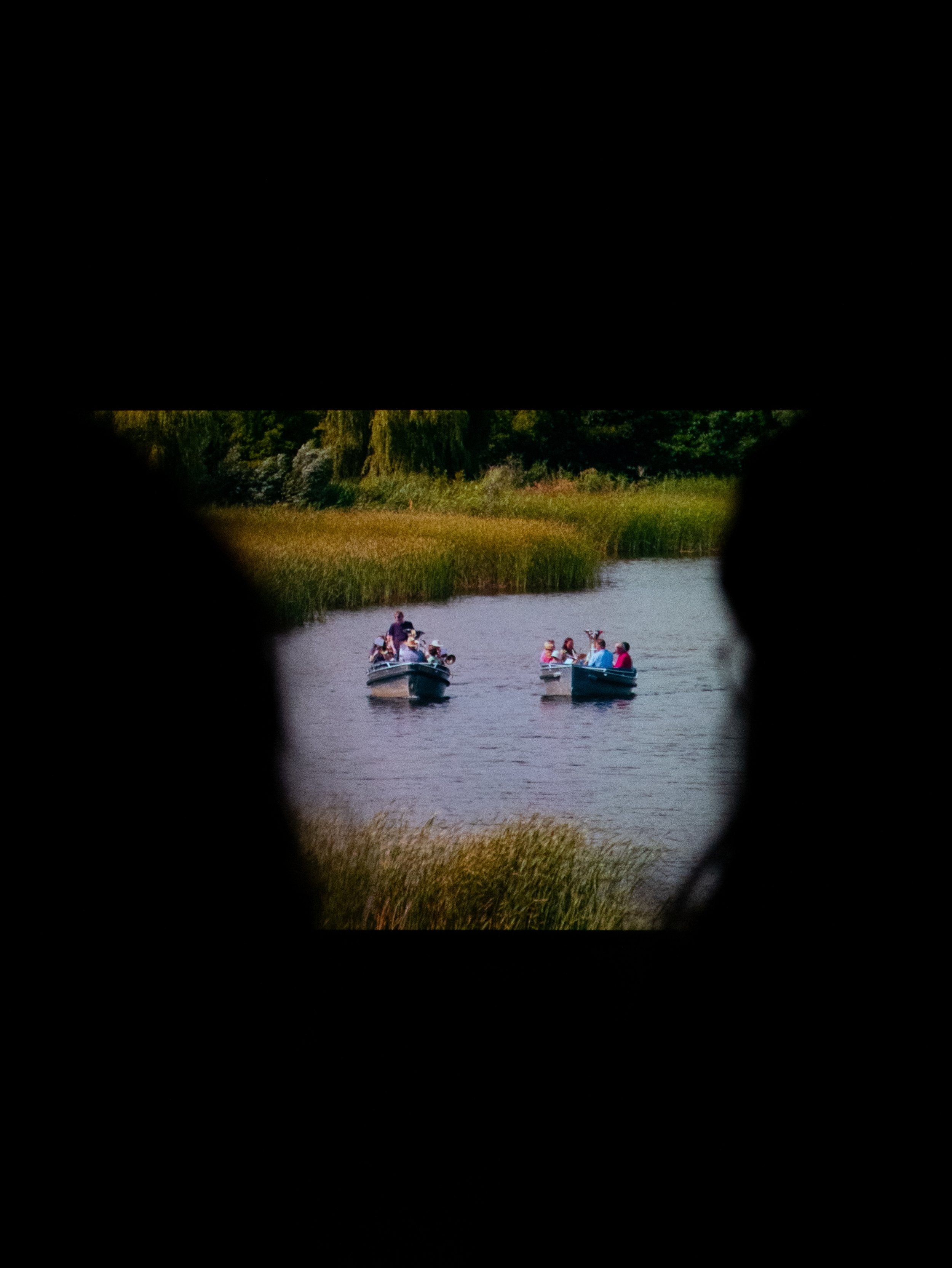 Looking through a narrow window at two boats with people on a lake, surrounded by green trees and tall grass.
