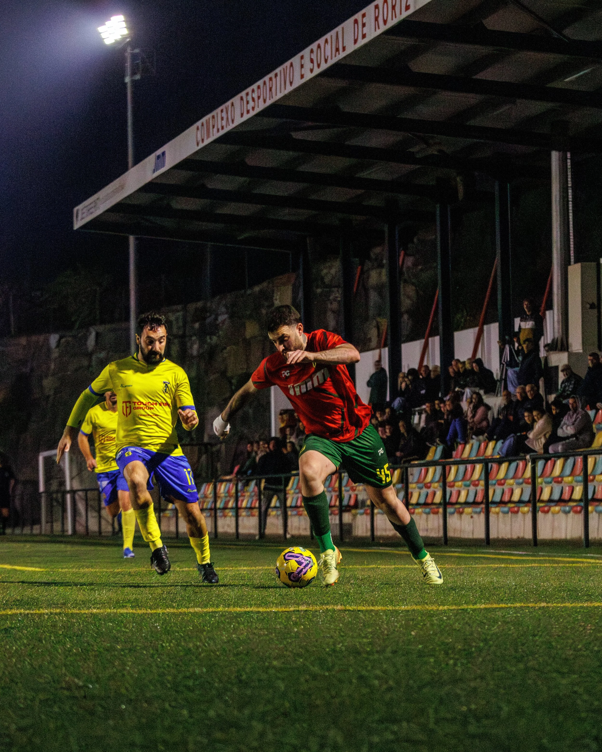 Soccer match occurring at night with players in yellow and green uniforms, and a player in red jersey controlling the ball on a well-lit field, with spectators watching from the bleachers.
