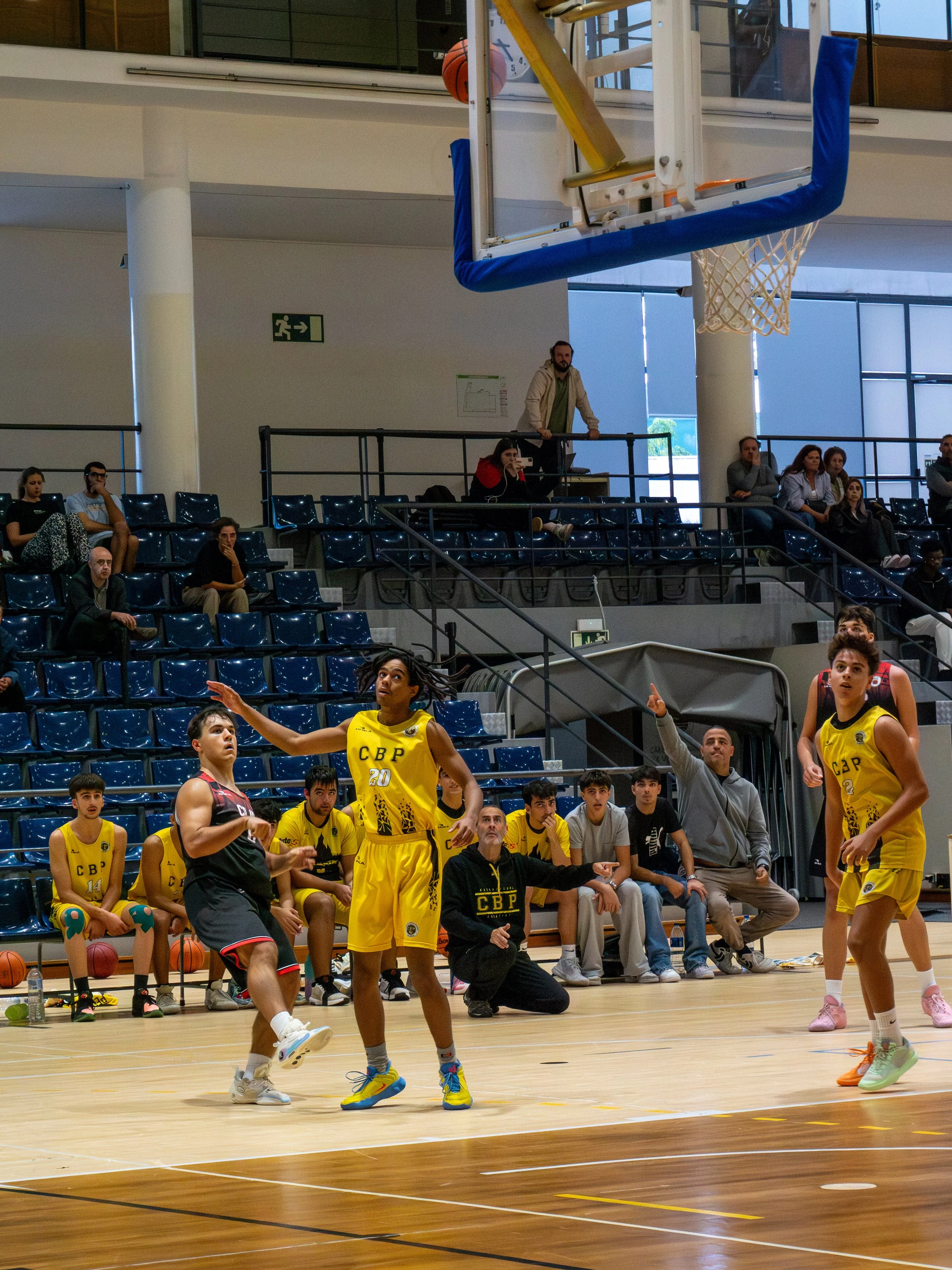 A youth basketball game in progress inside a gymnasium. Players in yellow and black jerseys are on the court, with some attempting to shoot or defend. Spectators and coaches watch from the bleachers and sidelines, while a basketball hoop hangs above.