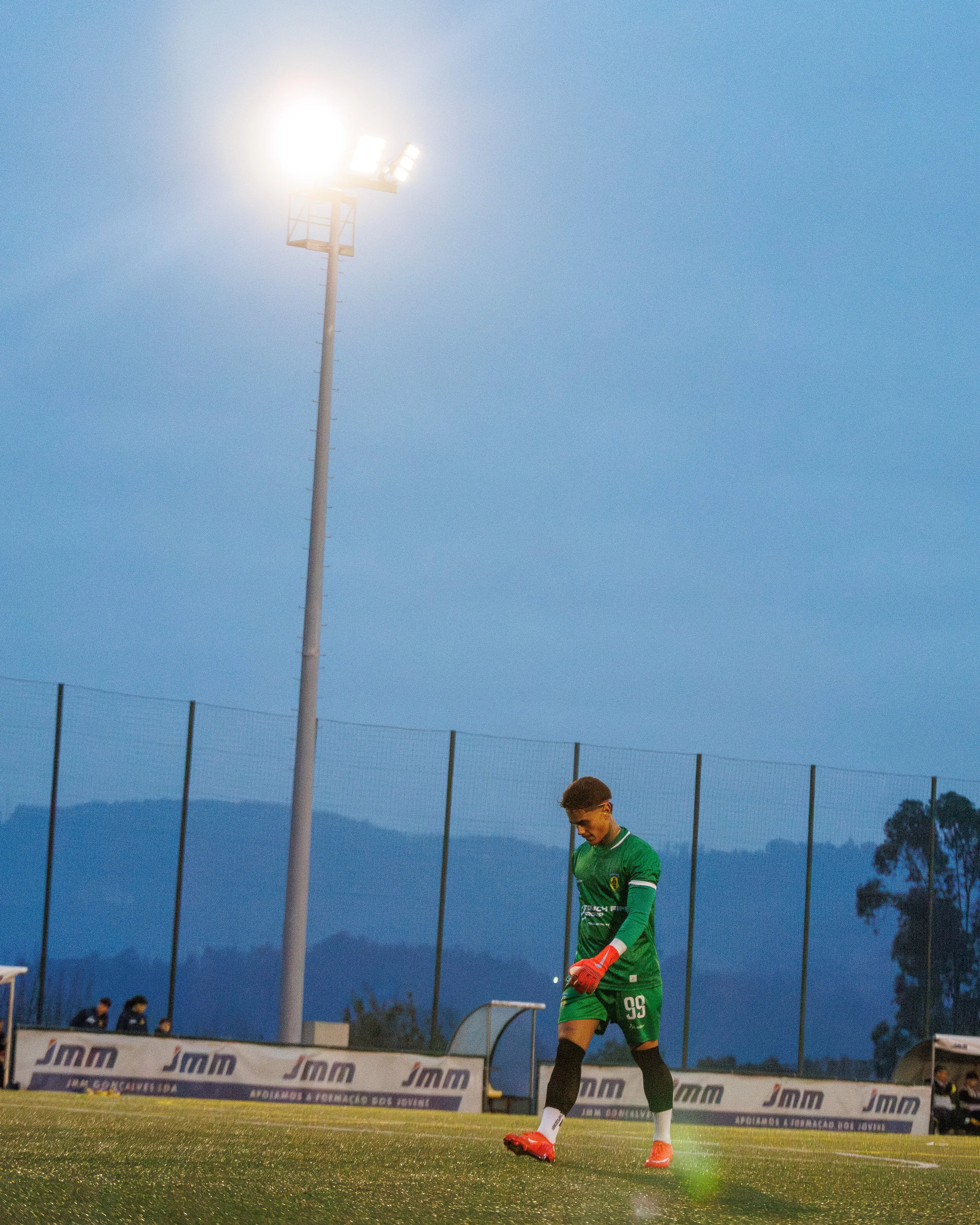 A soccer player dressed in a green uniform and red gloves walking on a field under stadium lights in the evening.