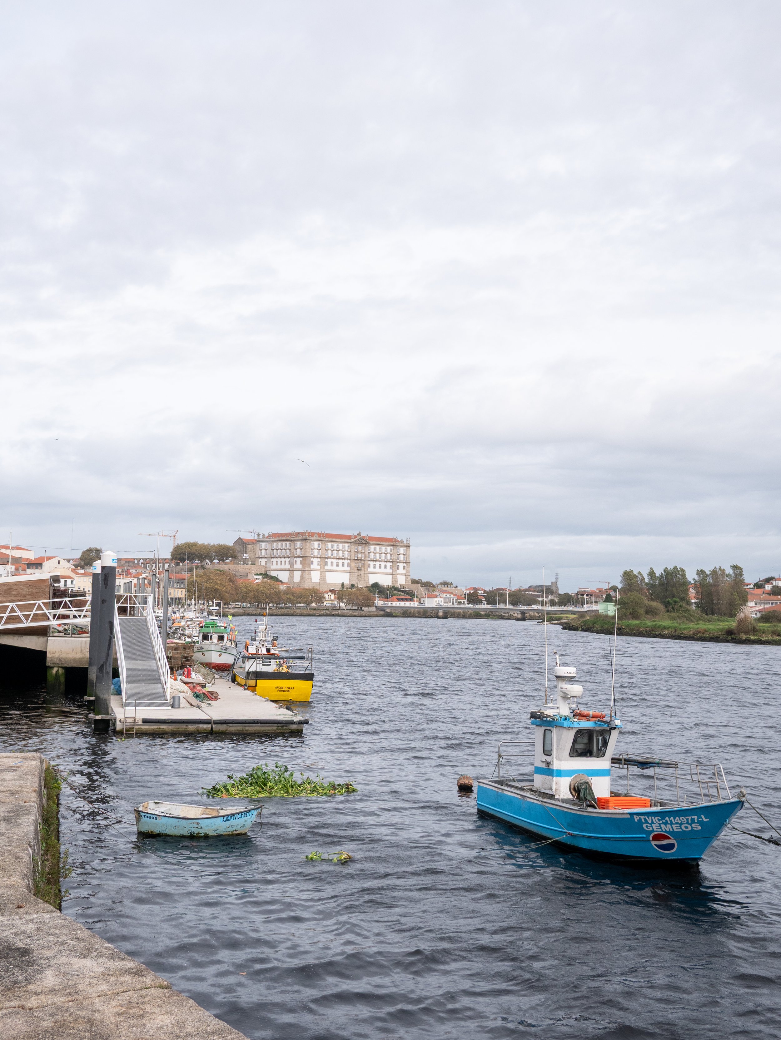 A harbor with boats docked along the pier, a castle-like building in the background, and a partly cloudy sky.