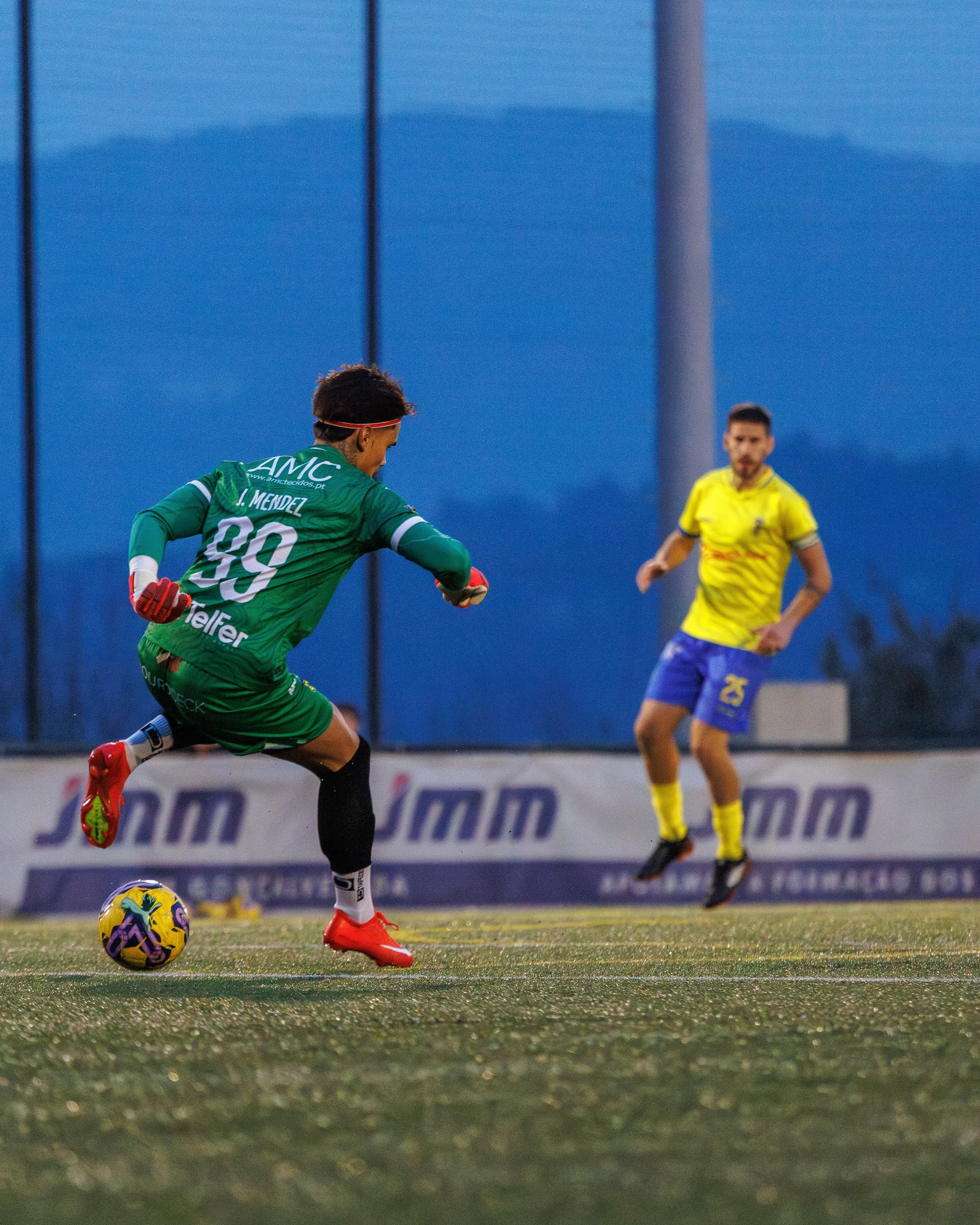 Soccer player in green jersey kicking a colorful ball during a game with an opposing player in yellow and blue in the background.