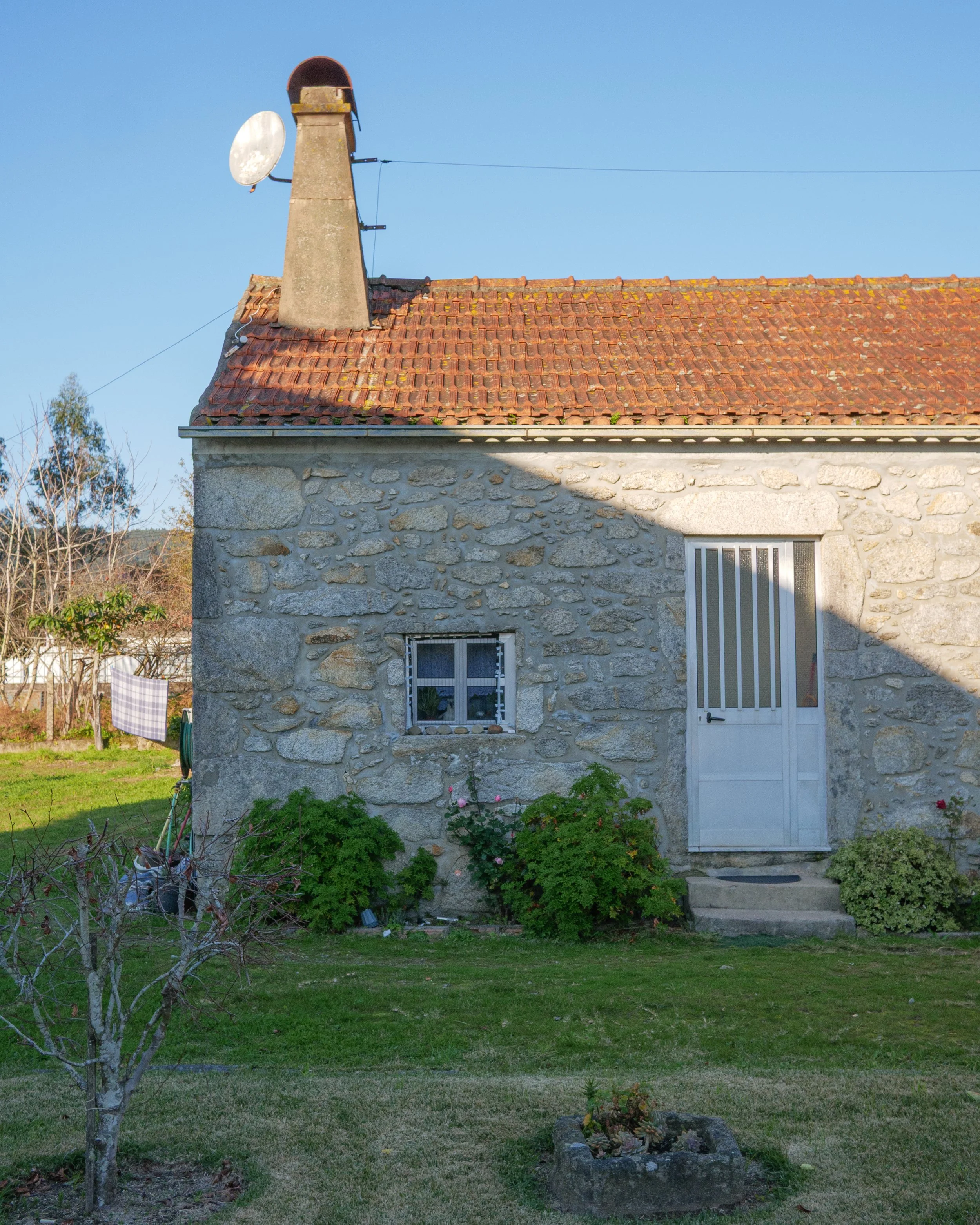 A stone house with a red tile roof, a small window, a white door, and a garden in front.