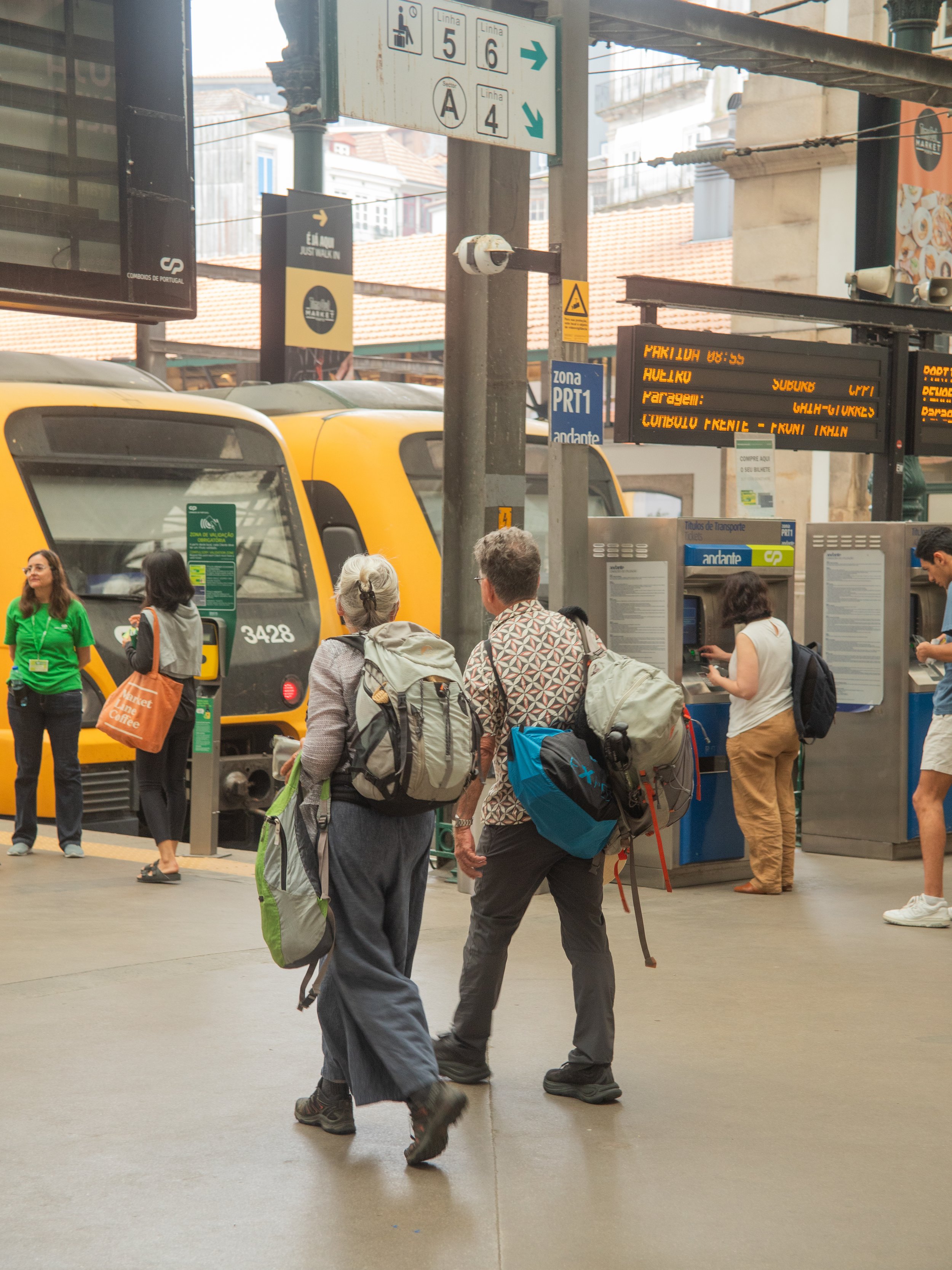 People with backpacks waiting at a train station, with yellow trains in the background and electronic signage showing departure information.