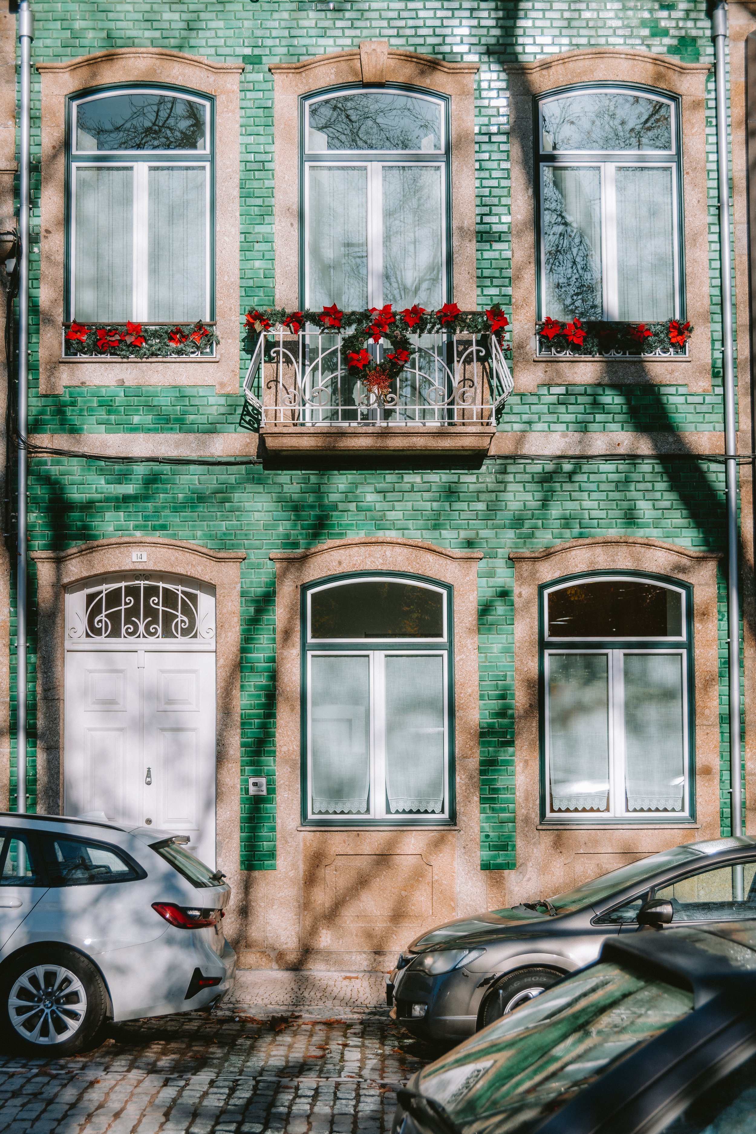 A green brick building with six large windows adorned with red holiday poinsettia decorated flower boxes. Two upper balcony windows have ornate white balconies with flowers, and two ones on ground level are behind white frames. In front of the buildi