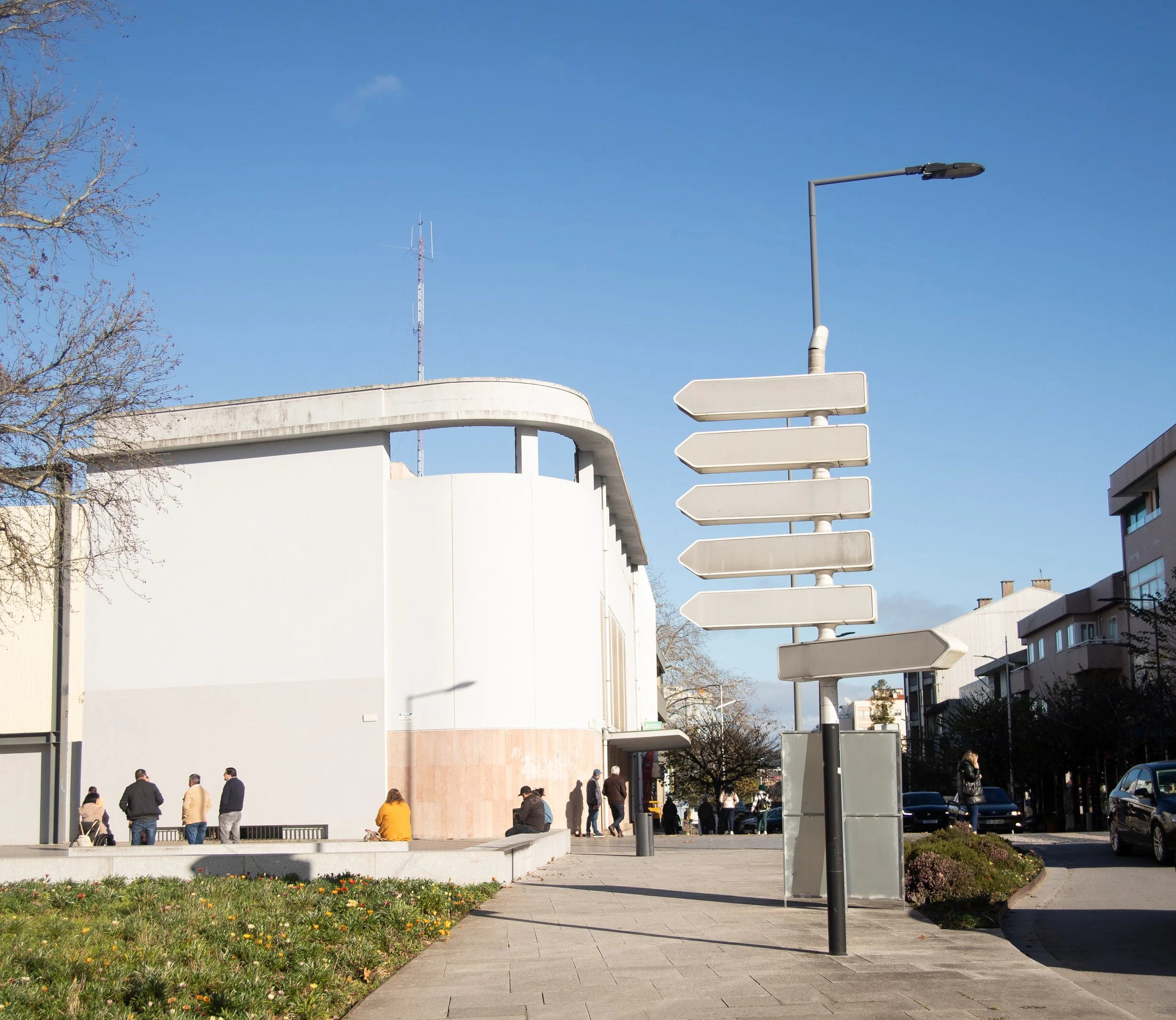 Street scene with a modern white building, people sitting and walking, a signpost with multiple blank directional signs, and a clear blue sky.