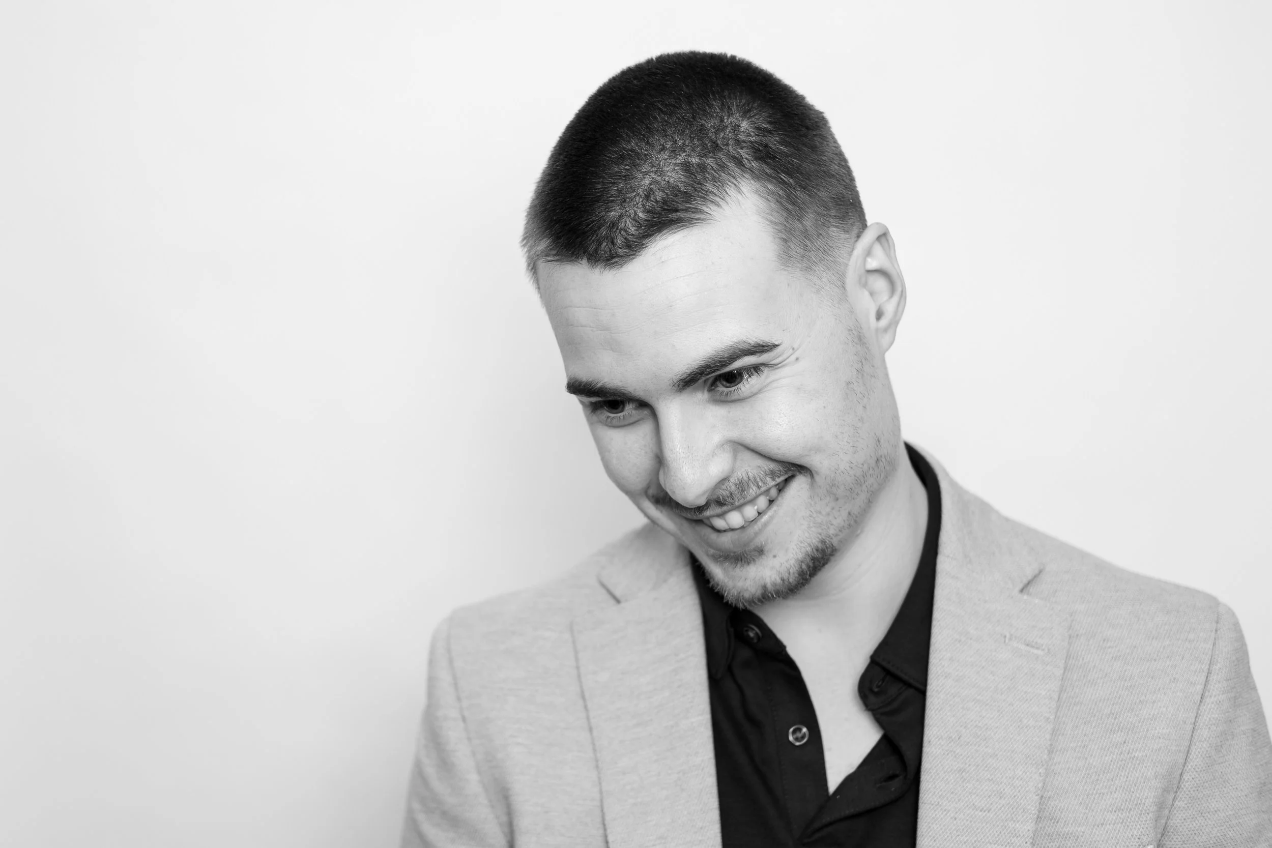 Black and white portrait of a smiling young man looking down, wearing a light blazer and a dark shirt, against a plain light background.