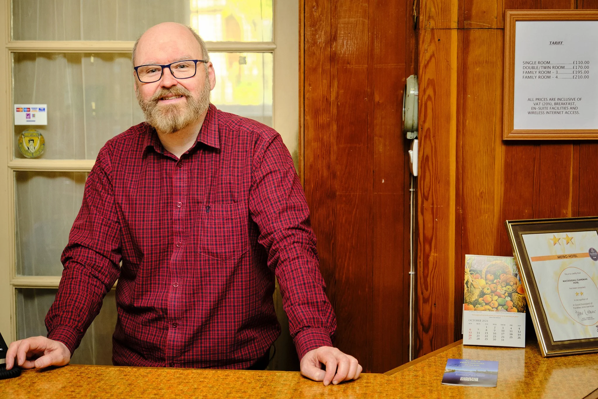 Man with glasses and a beard wearing a red plaid shirt standing behind a reception desk in a wooden-paneled office or hotel reception area.