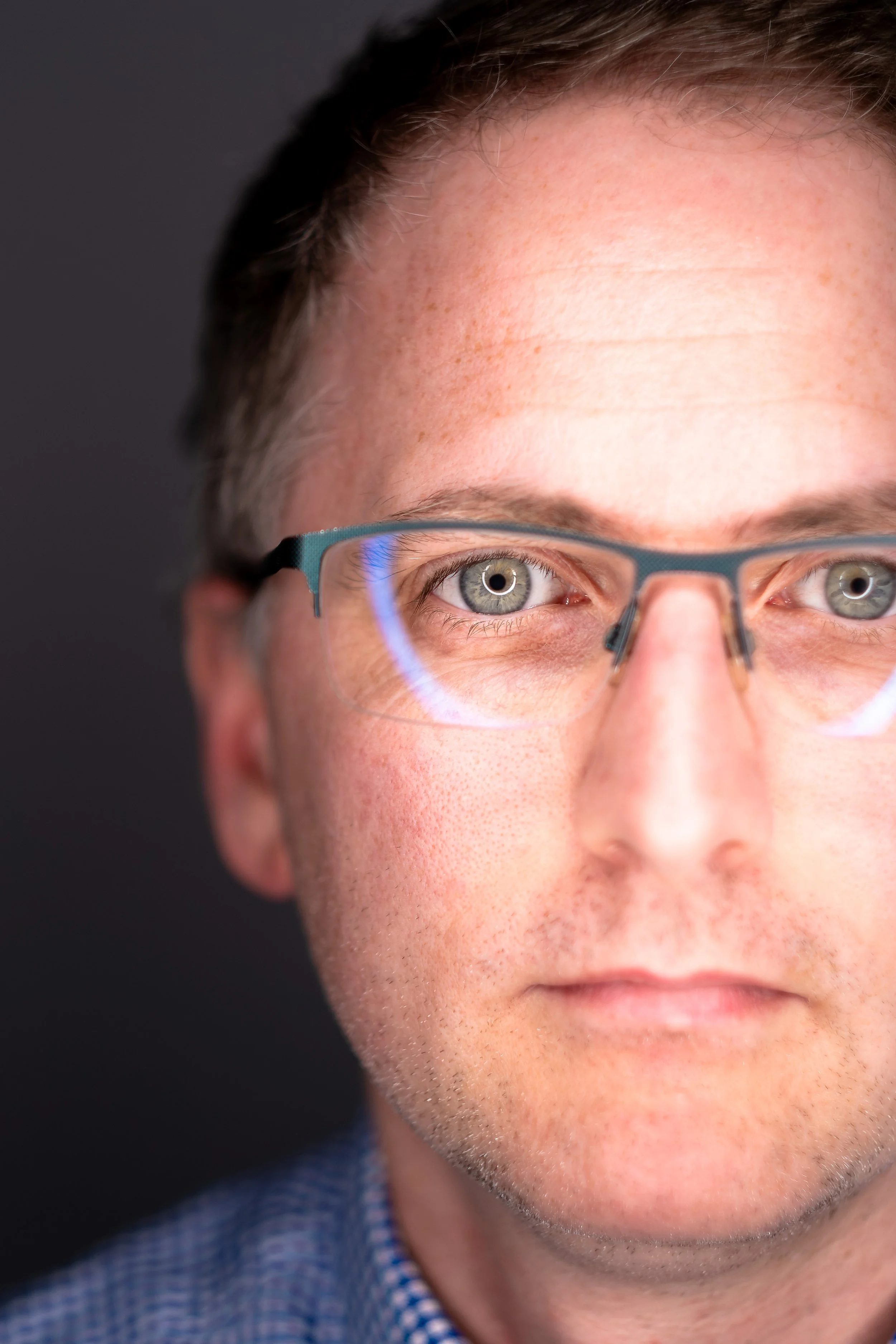 A close-up of a man with short hair and light skin wearing glasses, with a focused expression and a dark background.