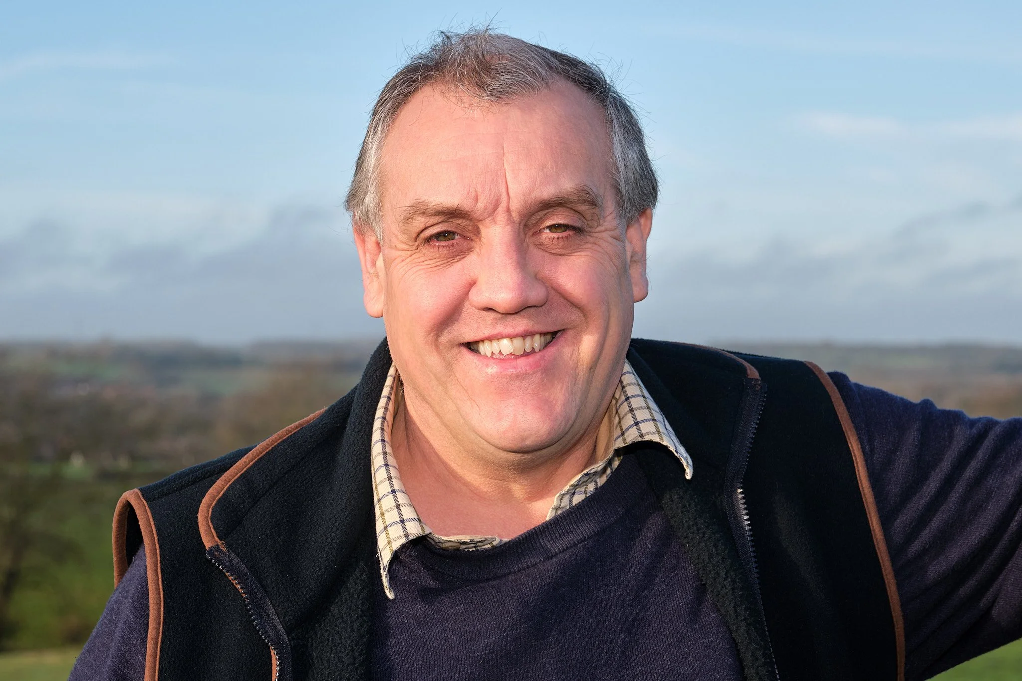 A smiling man with outdoors in front of a landscape with a cloudy sky.