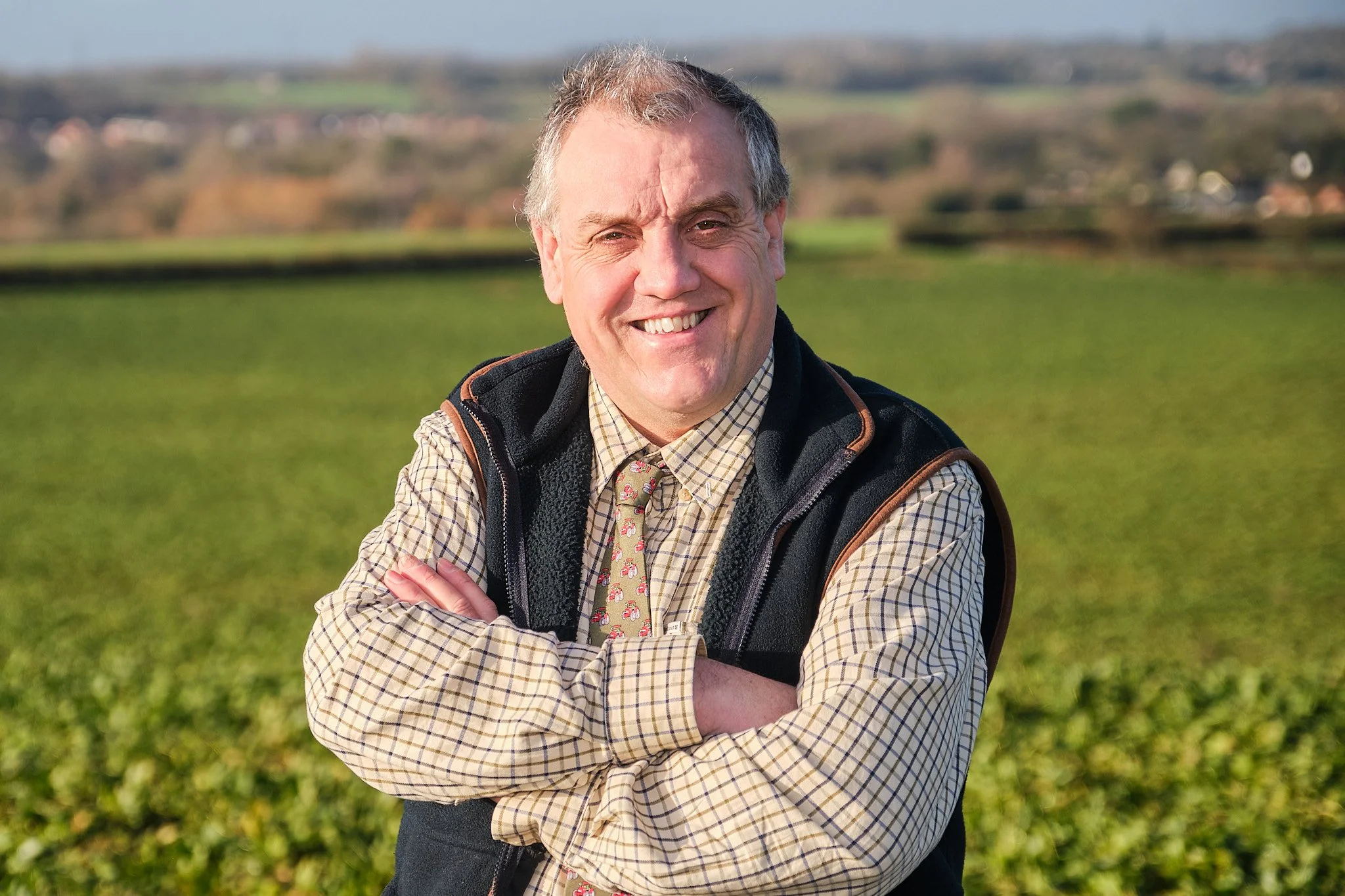 A smiling middle-aged man with gray hair, wearing a plaid shirt, a patterned tie, and a black vest with brown accents, stands outdoors in a green field with a blurred rural landscape in the background.