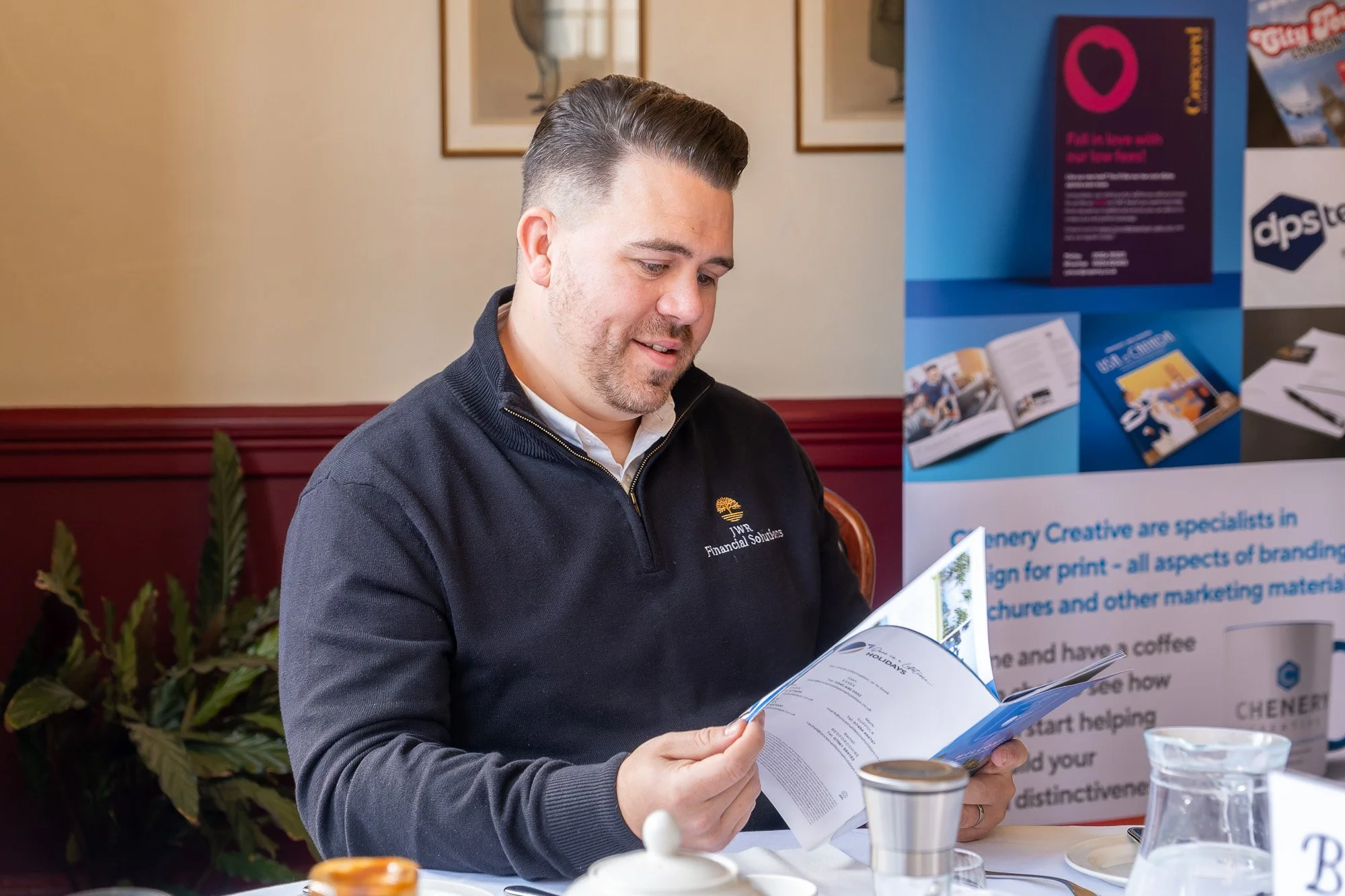 A man seated at a table, reading a brochure or booklet, with a banner and some informational materials in the background. He is wearing a dark zip-up sweater with a logo that reads 'JWR Financial Solutions'.