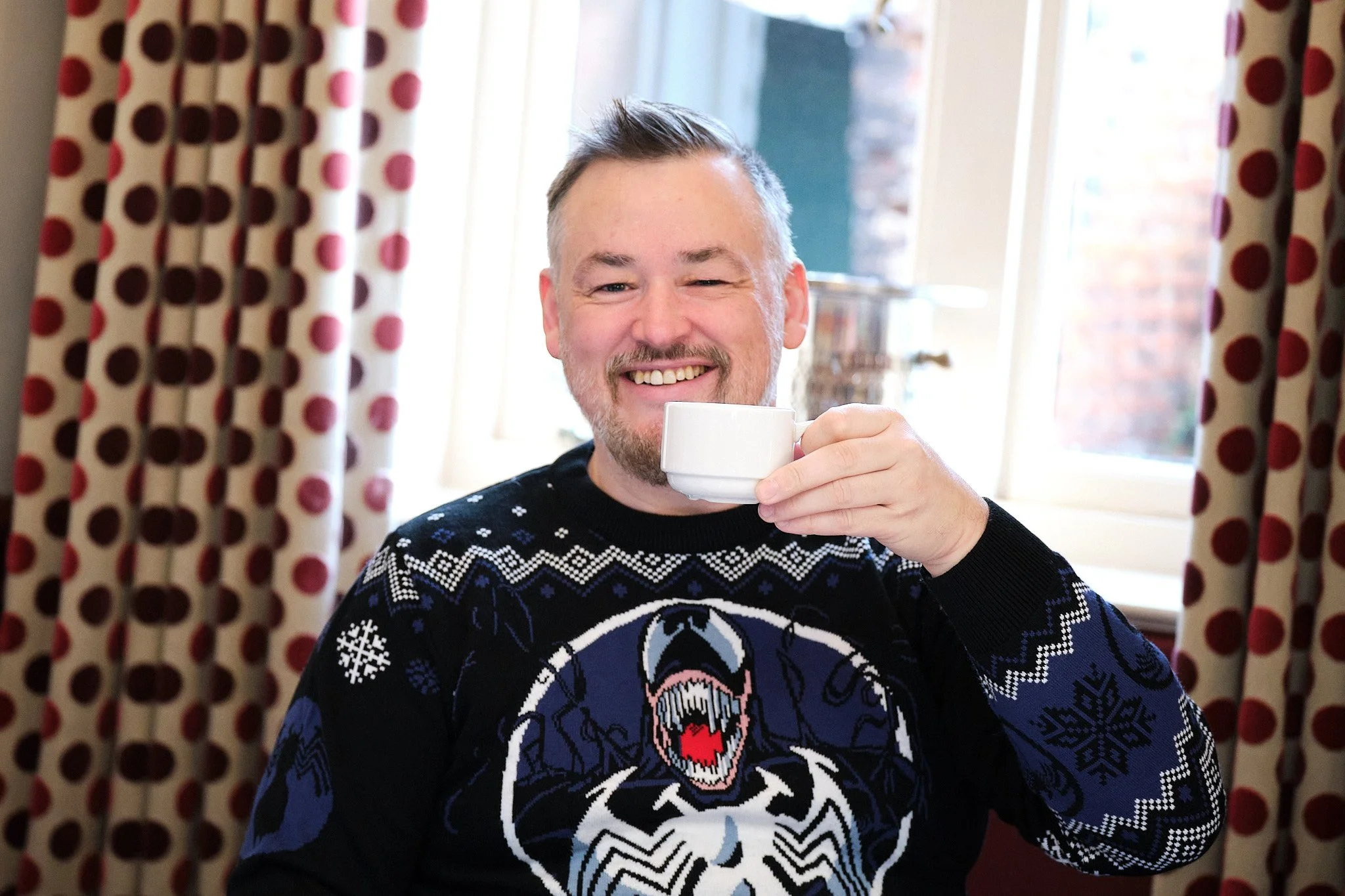 Man smiling and drinking from a white mug, wearing a black holiday sweater with a snowflake and skull design, sitting by a window with polka dot curtains.