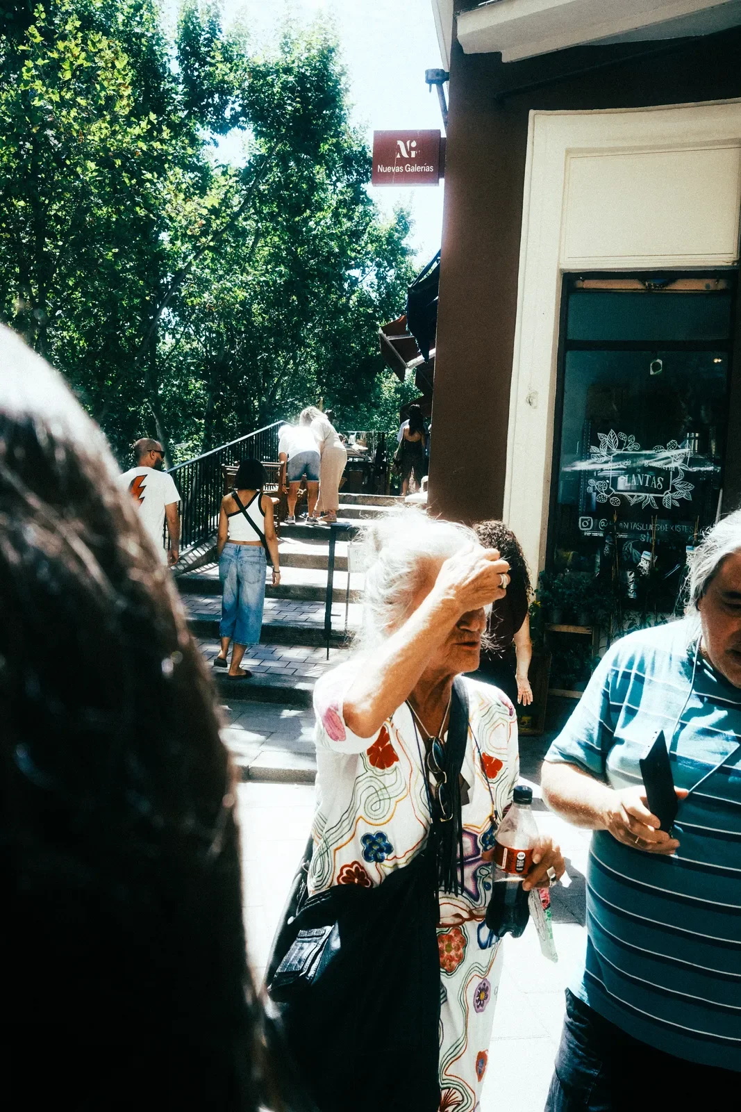 Varios peatones en una calle soleada con árboles, una mujer mayor con cabello gris y un vestido con diseño de flores, sosteniendo una botella y una bolsa negra.
