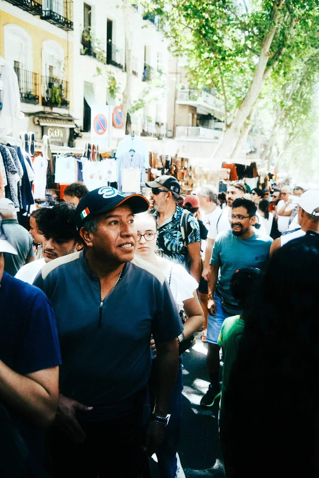 Imagen de una calle concurrida en un mercado al aire libre con numerosos puestos vendiendo ropa y accesorios, y muchas personas paseando bajo árboles en un día soleado en una ciudad.