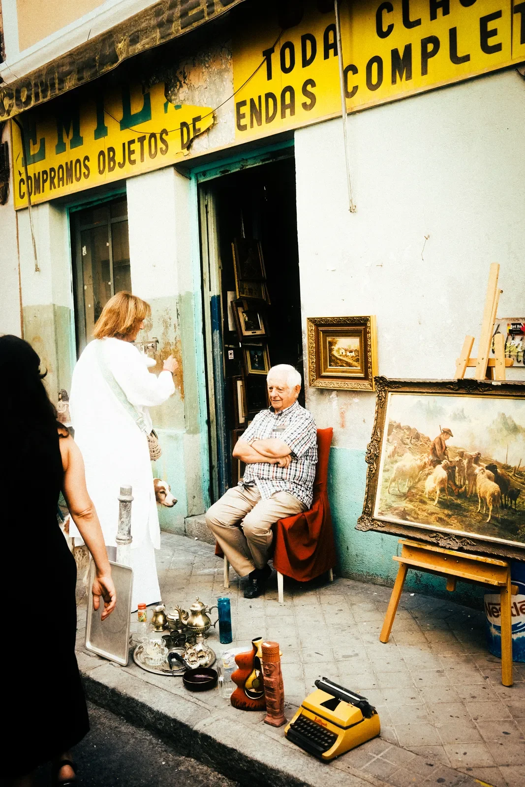 Un hombre sentado en una silla en la acera frente a una tienda de antigüedades exhibiendo objetos viejos y cuadros, con varias personas y un perro cerca, y un letrero en español que dice 'EMILDE Compraremos objetos de_...'.