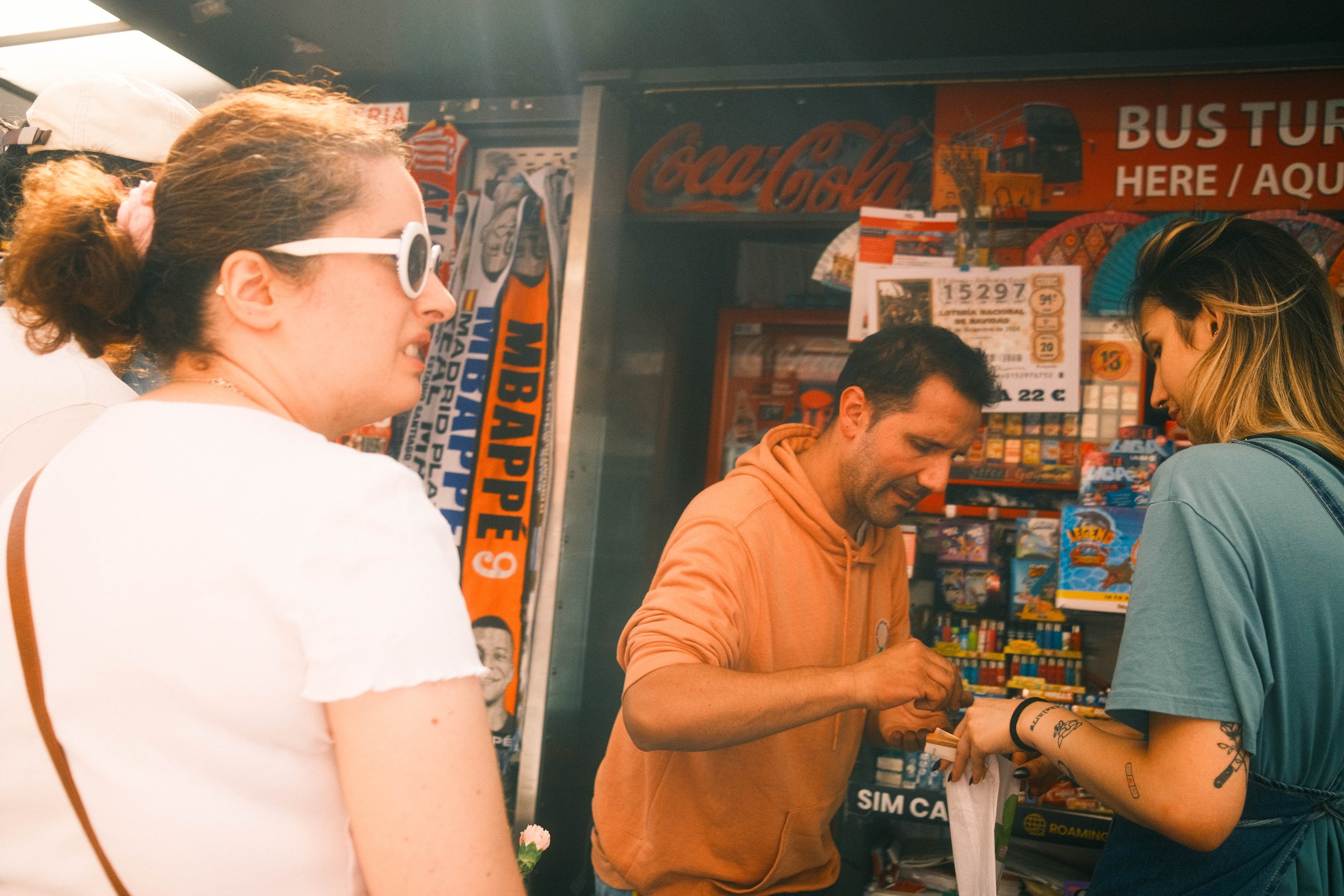 Una mujer con lentes blancos comprando algo en una tienda de souvenirs, atendida por un hombre y otra mujer con tatuajes