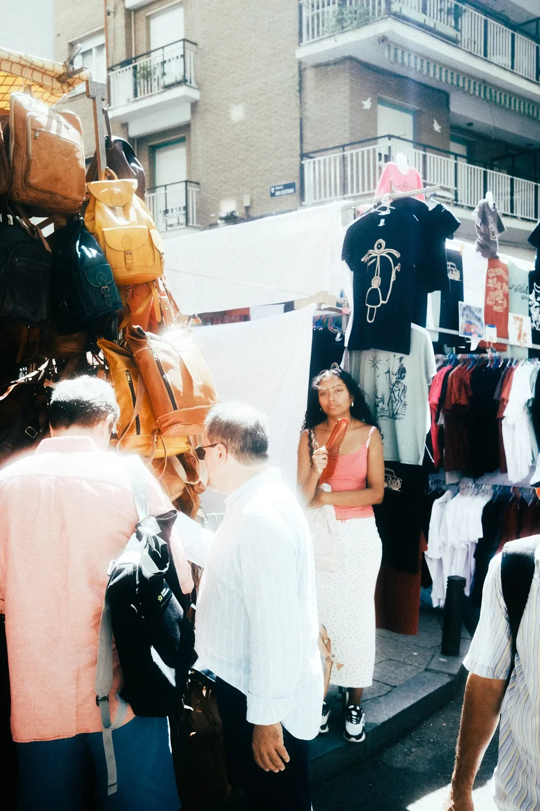 Una mujer de pie frente a un puesto de venta en la calle, sosteniendo un artículo en la mano; al fondo y a su alrededor, hay varias personas y ropa en exhibición, con edificios de apartamentos en el fondo.