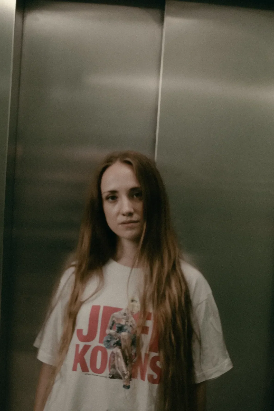 Joven mujer con cabello largo y rizado, con camiseta blanca de diseño y texto en rojo, en un ascensor de paredes metálicas.