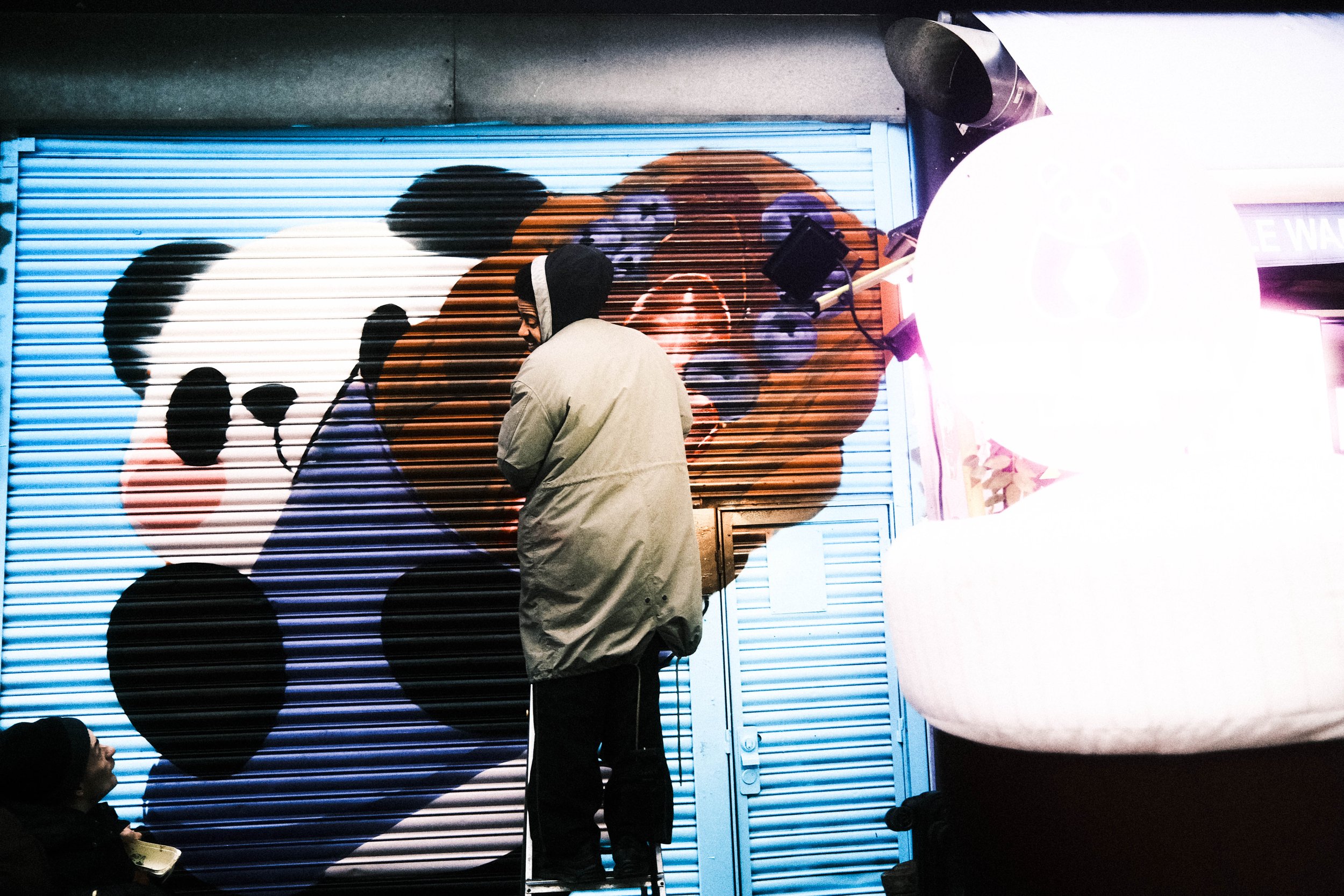 Persona pintando un mural de panda y oso de peluche en una persiana metálica en la calle.