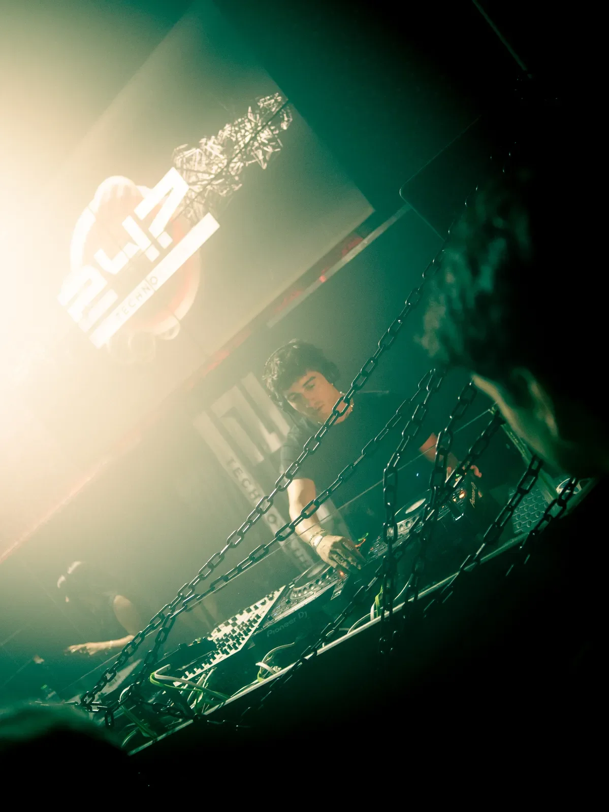 DJ mezclando en un club nocturno con iluminación de neón y cadenas en primer plano.